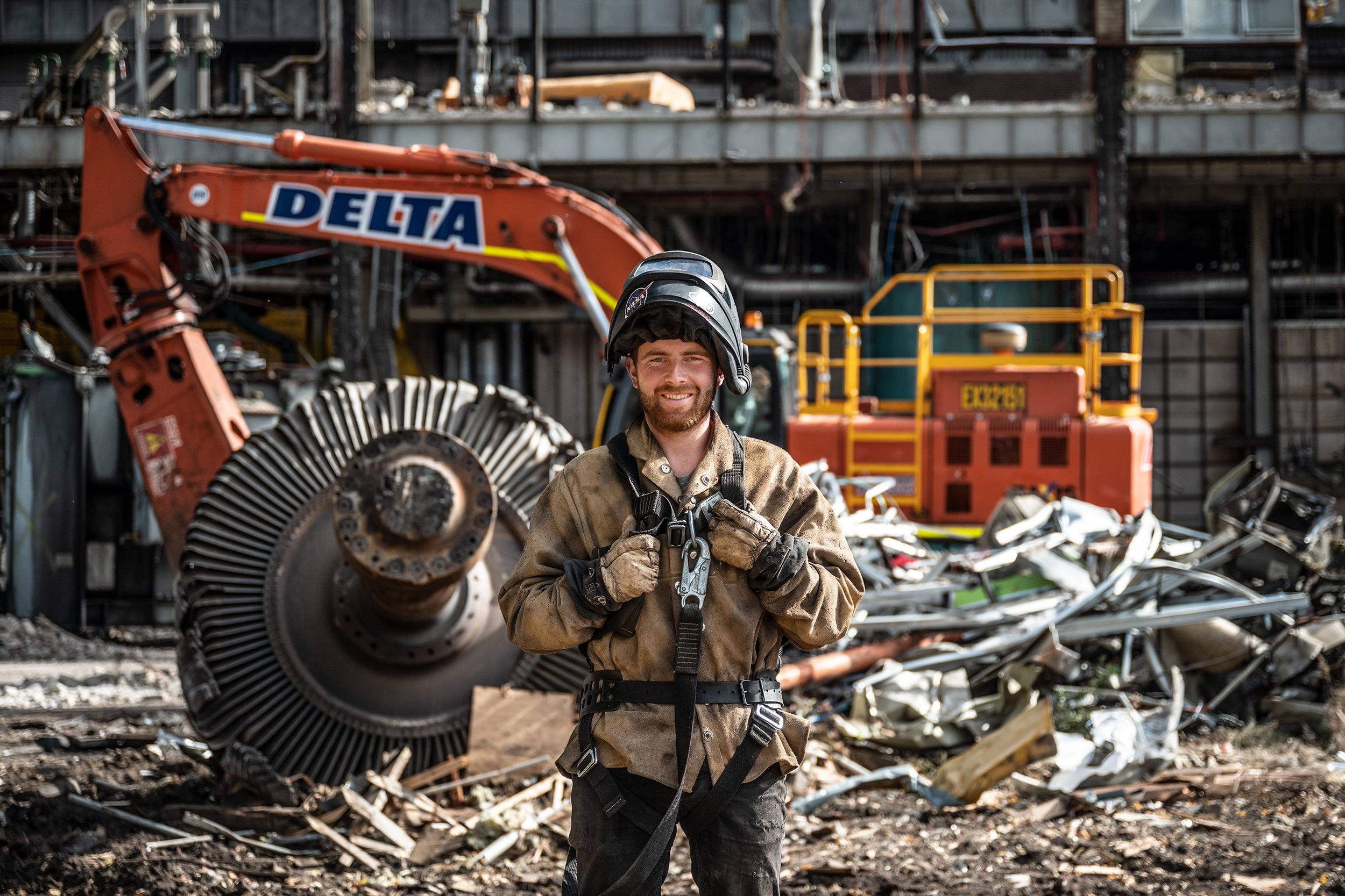 A firefighter in work gear and helmet standing in front of wreckage with construction equipment and debris at a demolition site.