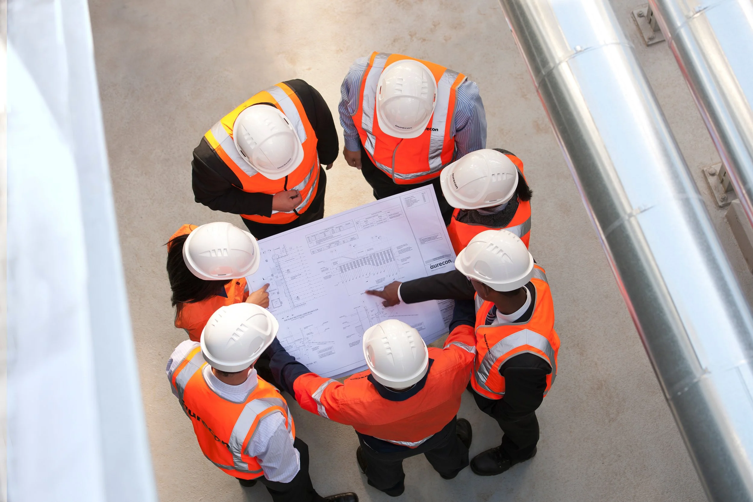 Group of construction workers wearing safety helmets and orange safety vests gathered around a large blueprint or plan, discussing it at a construction site.