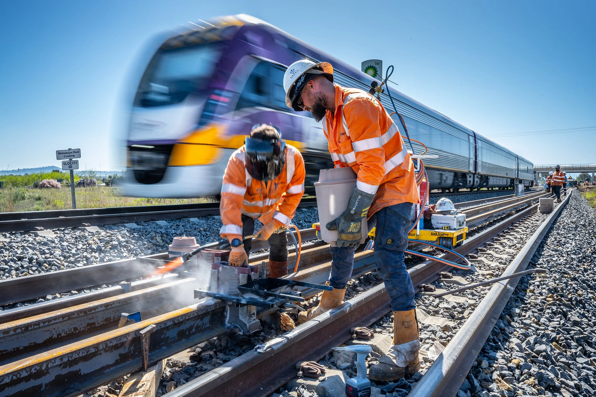 Two railway workers in orange safety uniforms and helmets working on a railway track, with a moving purple and yellow train passing in the background.