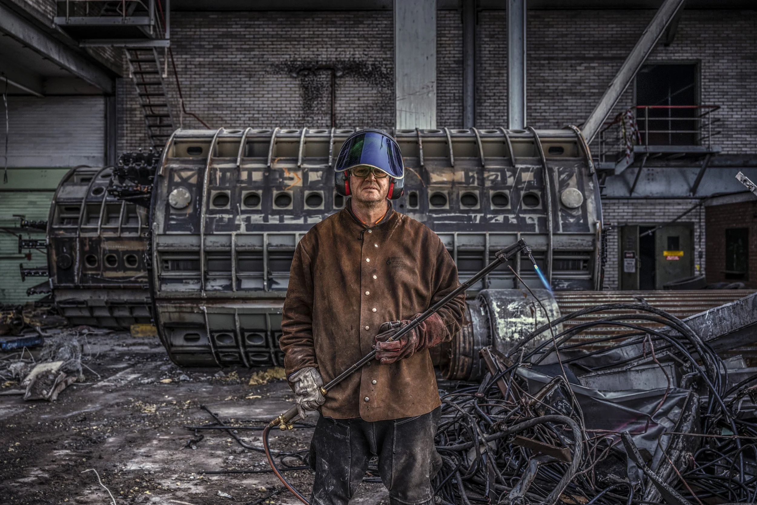 A man wearing protective gear, including a face shield, headphones, gloves, and a brown jacket, holding a torch in an industrial junkyard surrounded by scrap metal and debris.