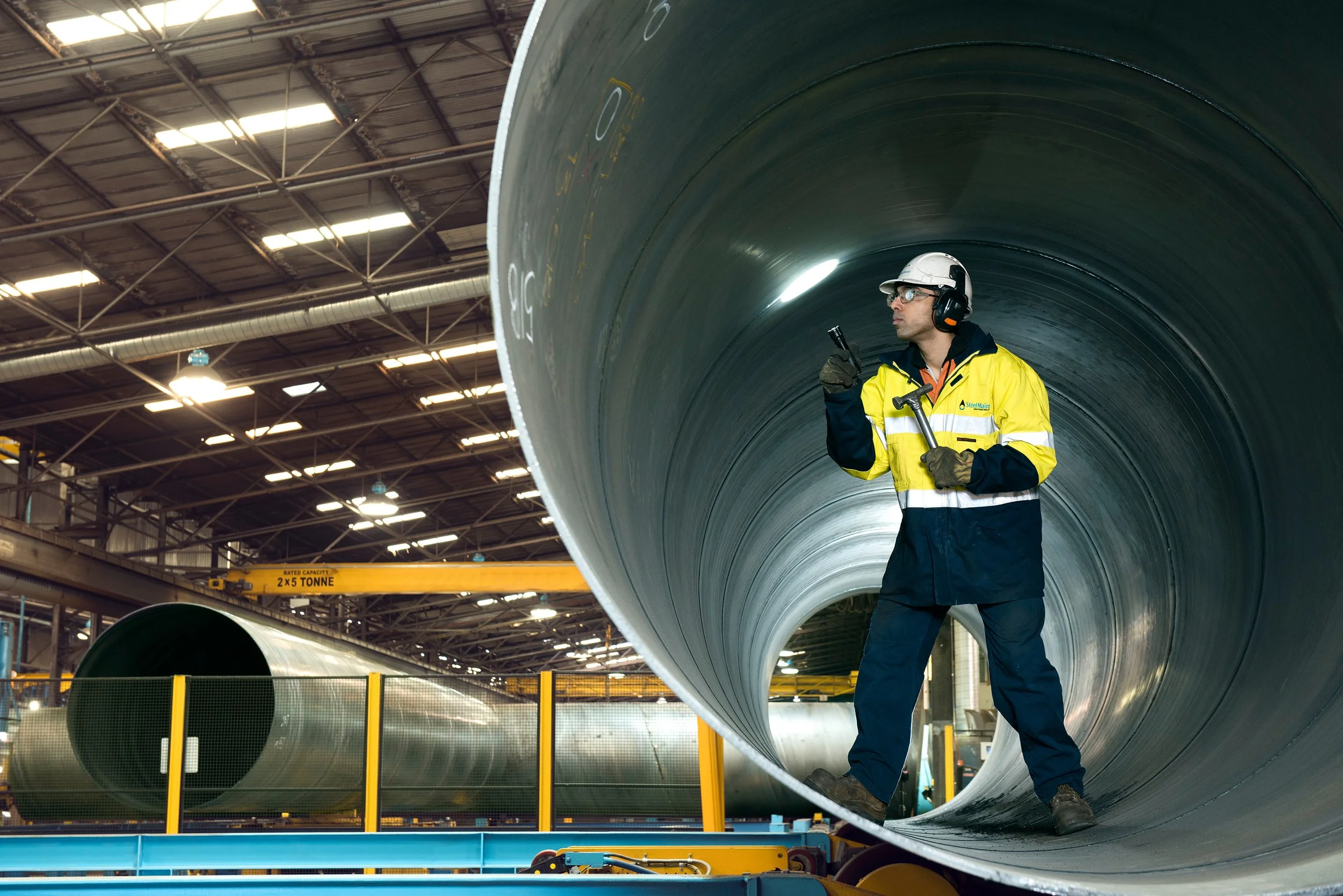 A worker in safety gear standing inside a large industrial pipe in a factory or warehouse setting.