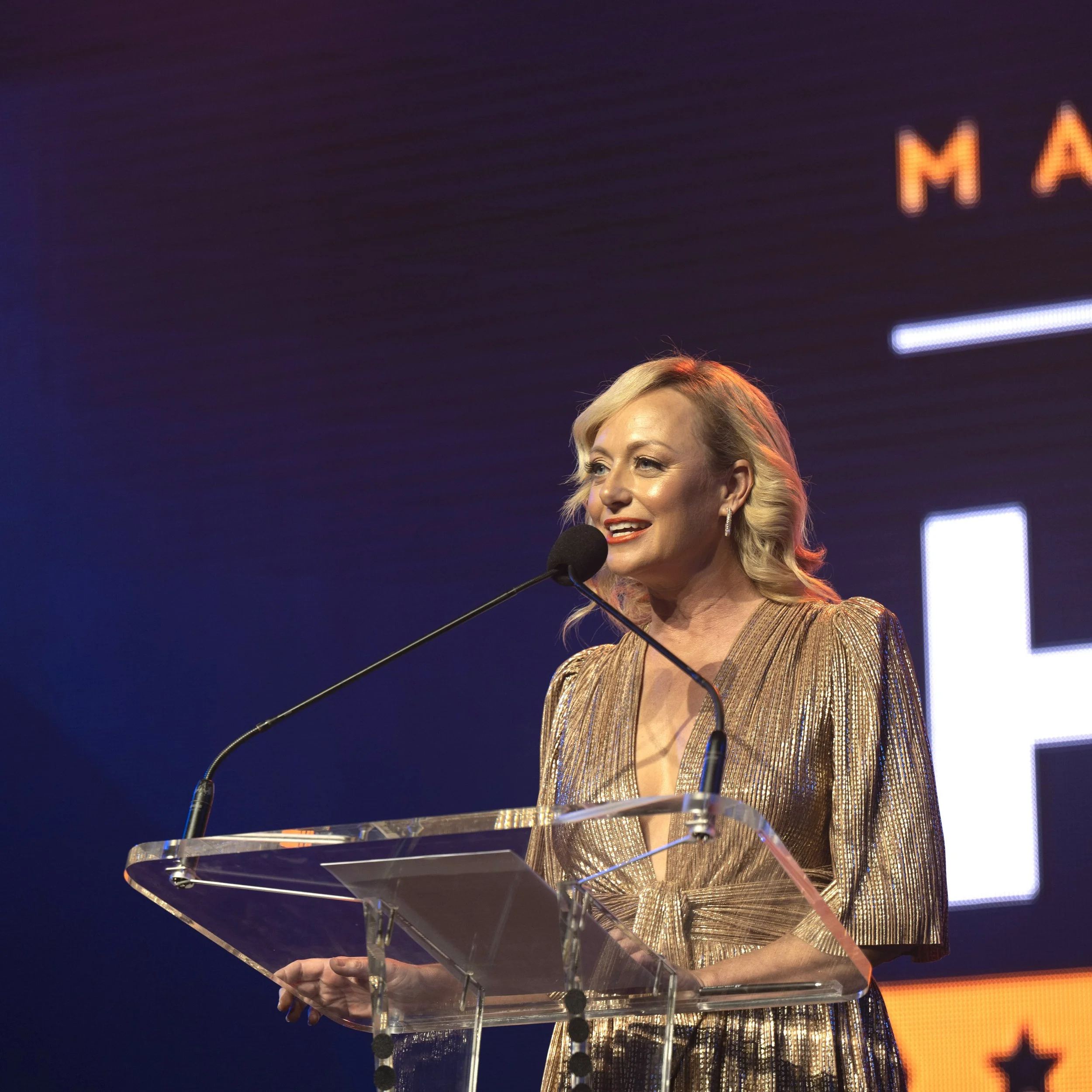 A woman with blonde hair in a gold dress speaking at a podium with a microphone on stage at an event.