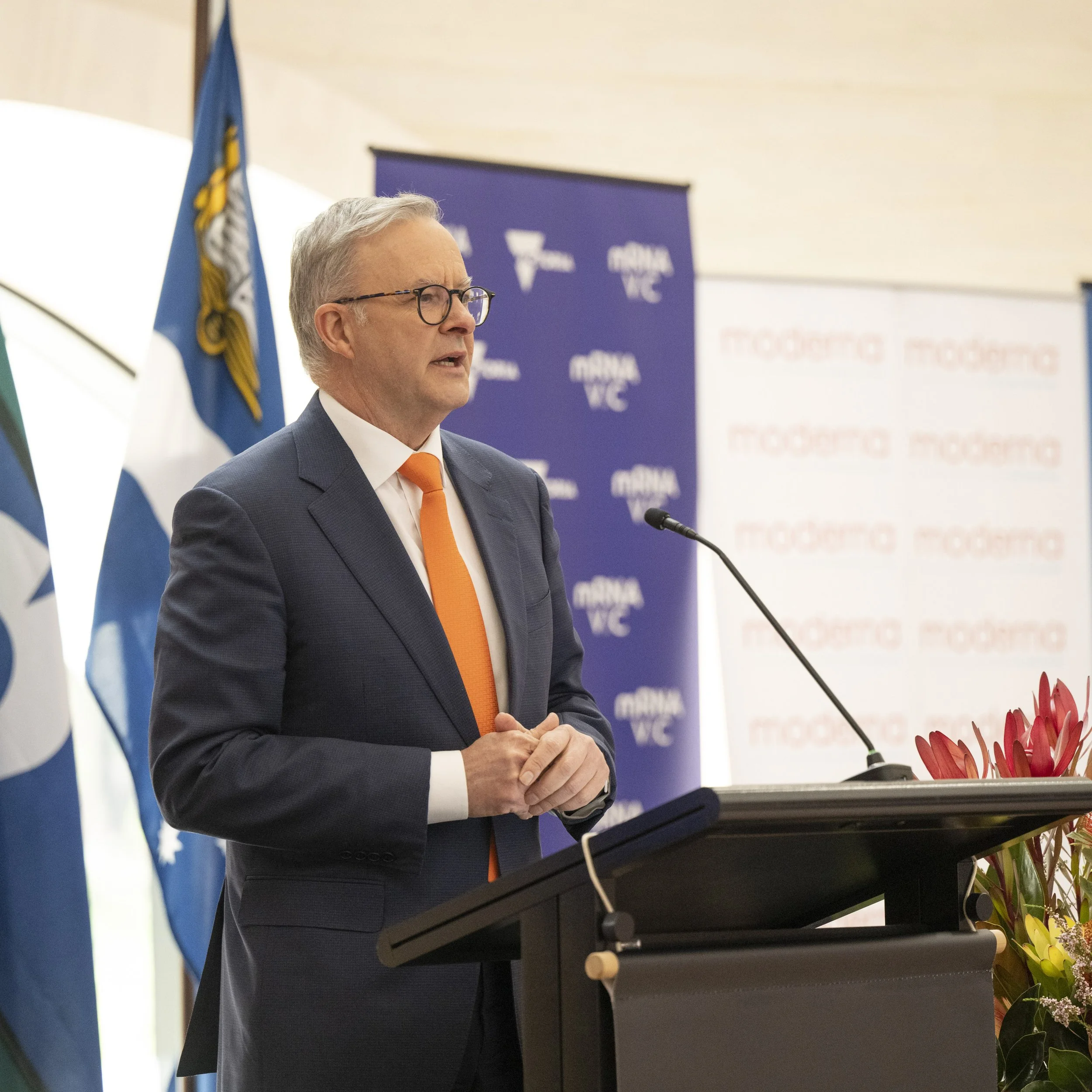 A man in a suit and orange tie speaking at a podium, with flags and a backdrop displaying 'MINA VC' and 'PRODUCING' in the background.