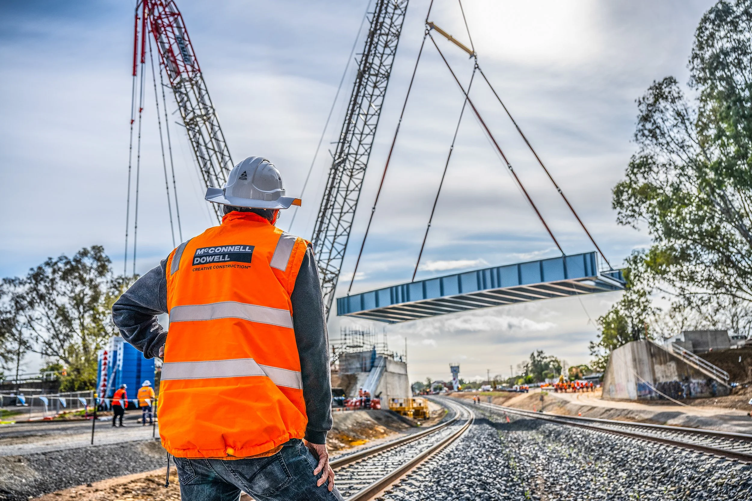 A construction worker in an orange safety vest and a white helmet watching a crane lift a section of a bridge over railroad tracks at a construction site.