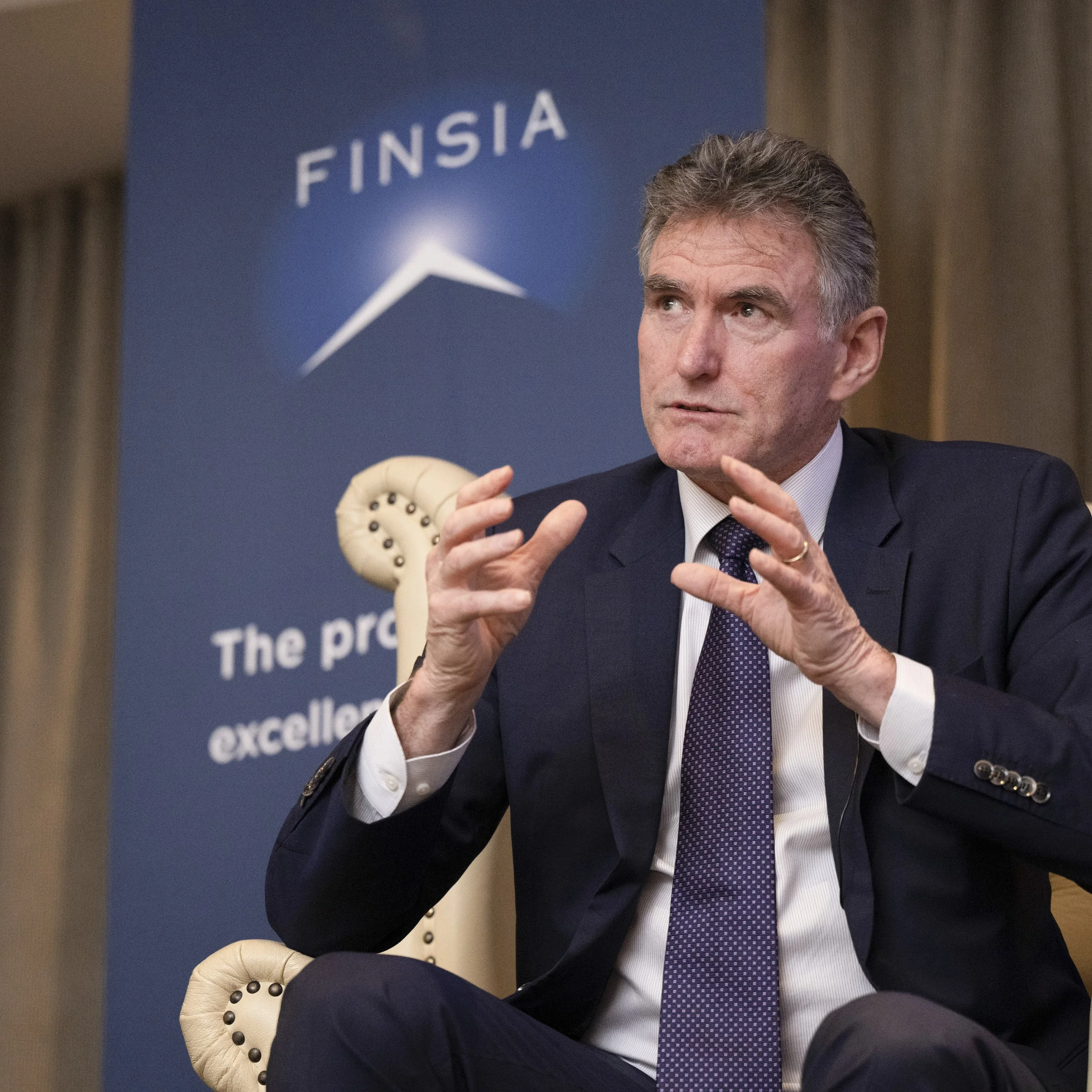 A man in a suit sitting in front of a blue banner with the word 'FINSIA' and a logo, speaking during a formal event.