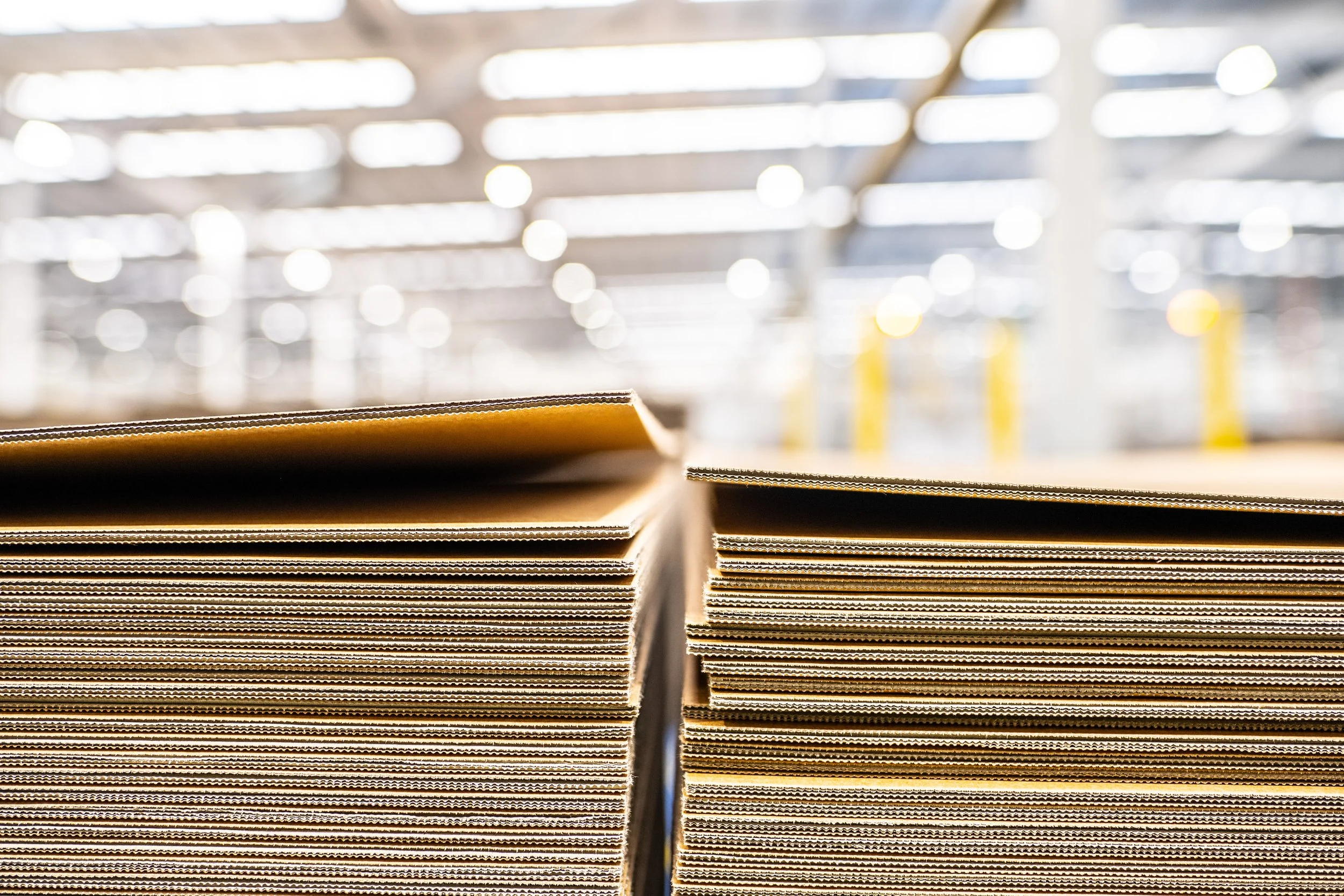 Close-up of stacked cardboard boxes or flat cardboard sheets in a warehouse or storage facility with blurred background and bright overhead lights.