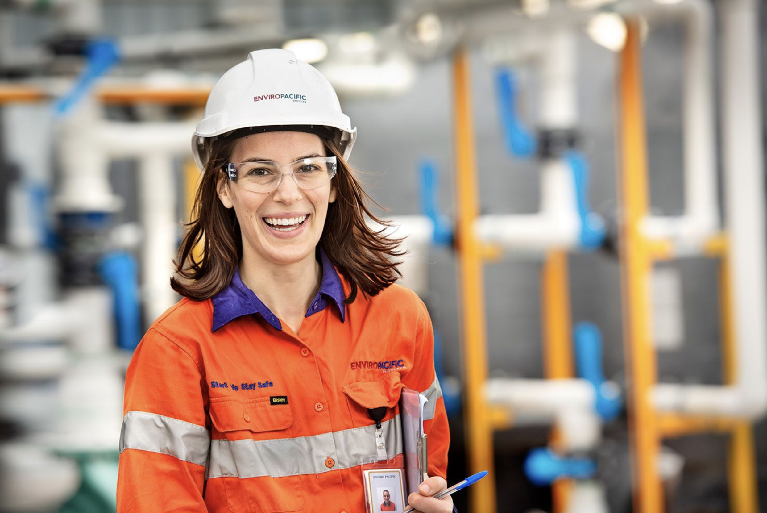Smiling woman in safety gear at industrial facility, wearing orange safety jacket, white helmet, and clear safety glasses, holding a pen and ID badge.