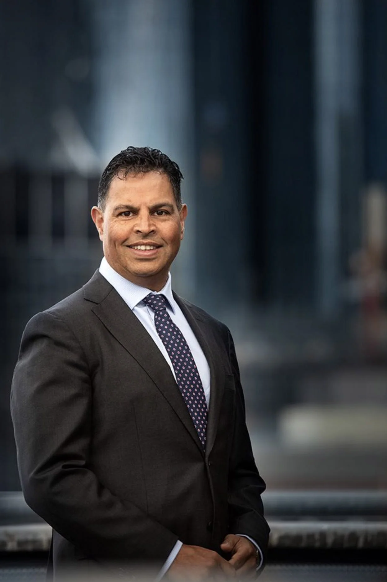 A man in a suit standing outdoors against a blurred cityscape background.