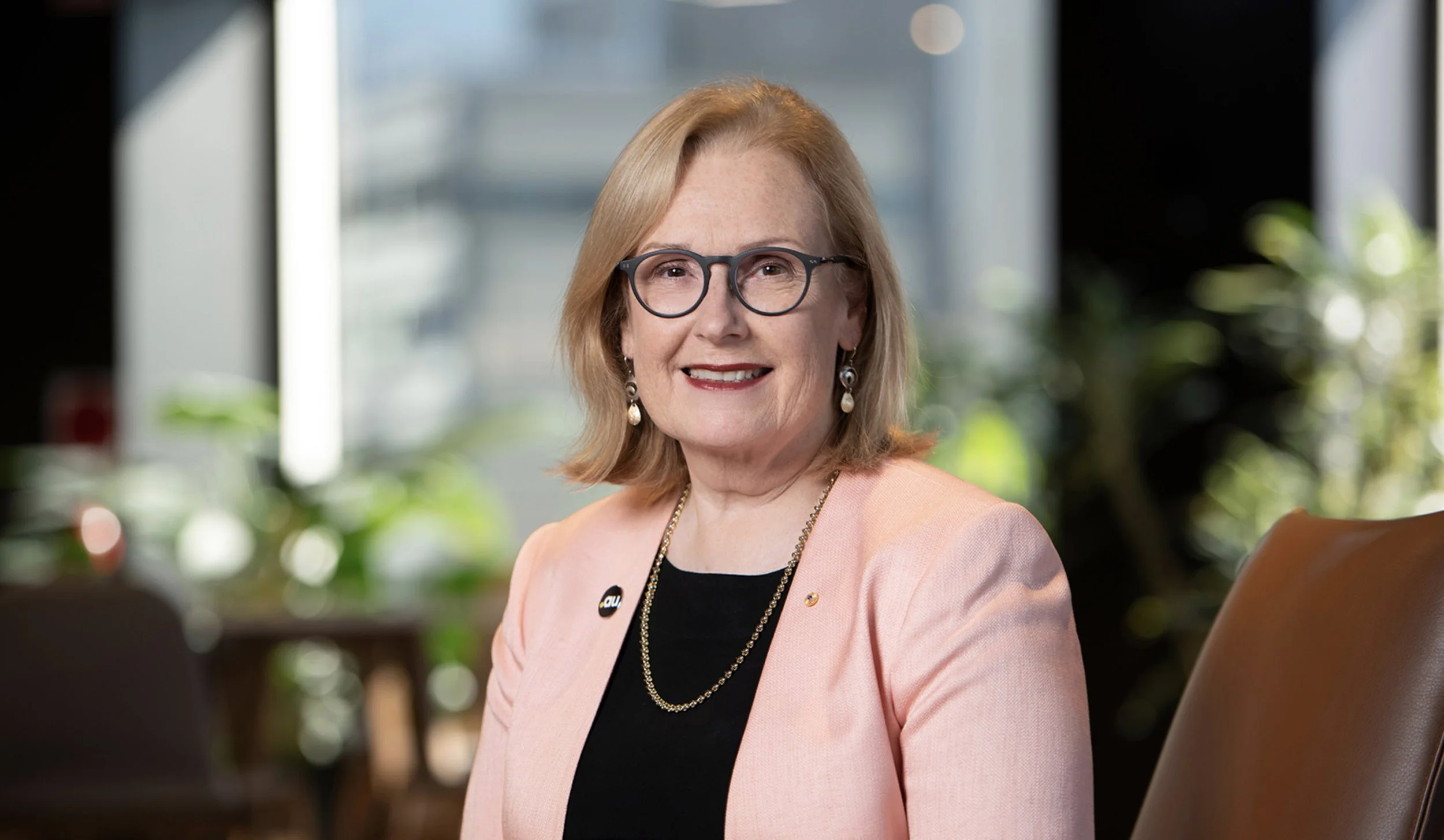 A professional woman with shoulder-length blonde hair, wearing glasses, pearl earrings, a black top, and a light pink blazer, sitting in a well-lit office environment.