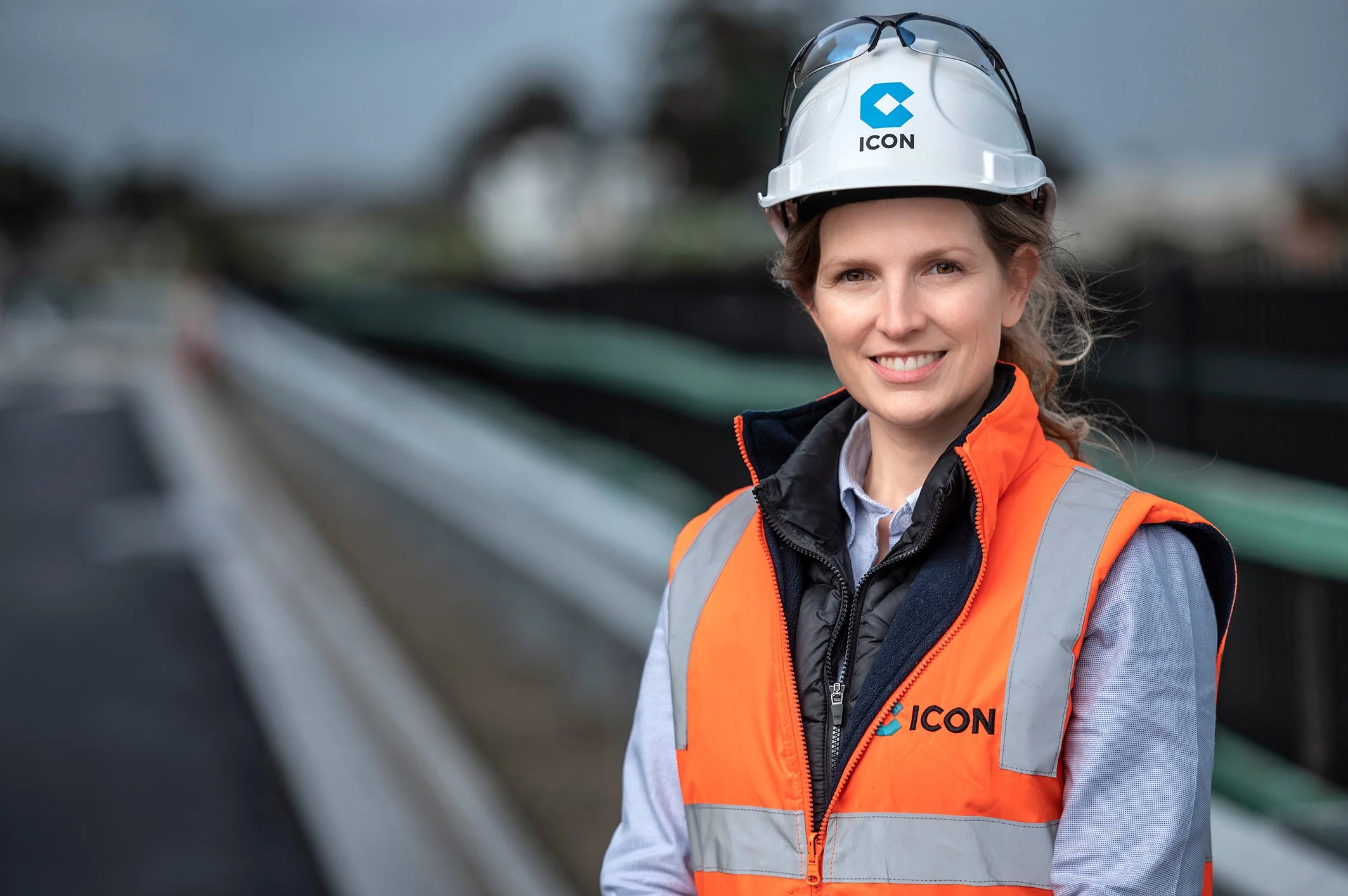 A smiling woman wearing a white safety helmet with sunglasses on top, an orange reflective safety vest with gray stripes, and a dark jacket underneath, standing outdoors near construction or industrial site.