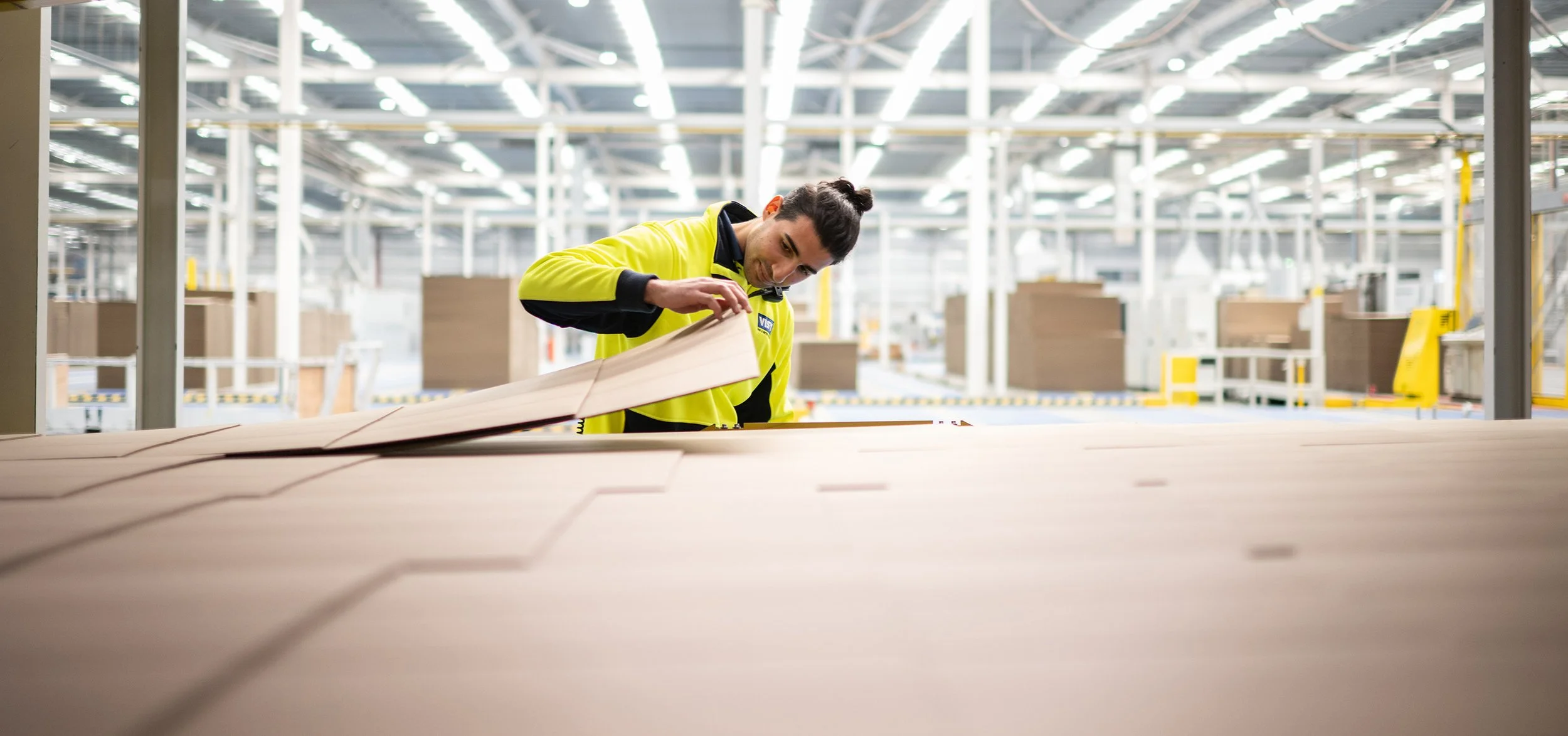 A man inspecting wooden boards in a warehouse or manufacturing facility, wearing a yellow jacket with black accents.