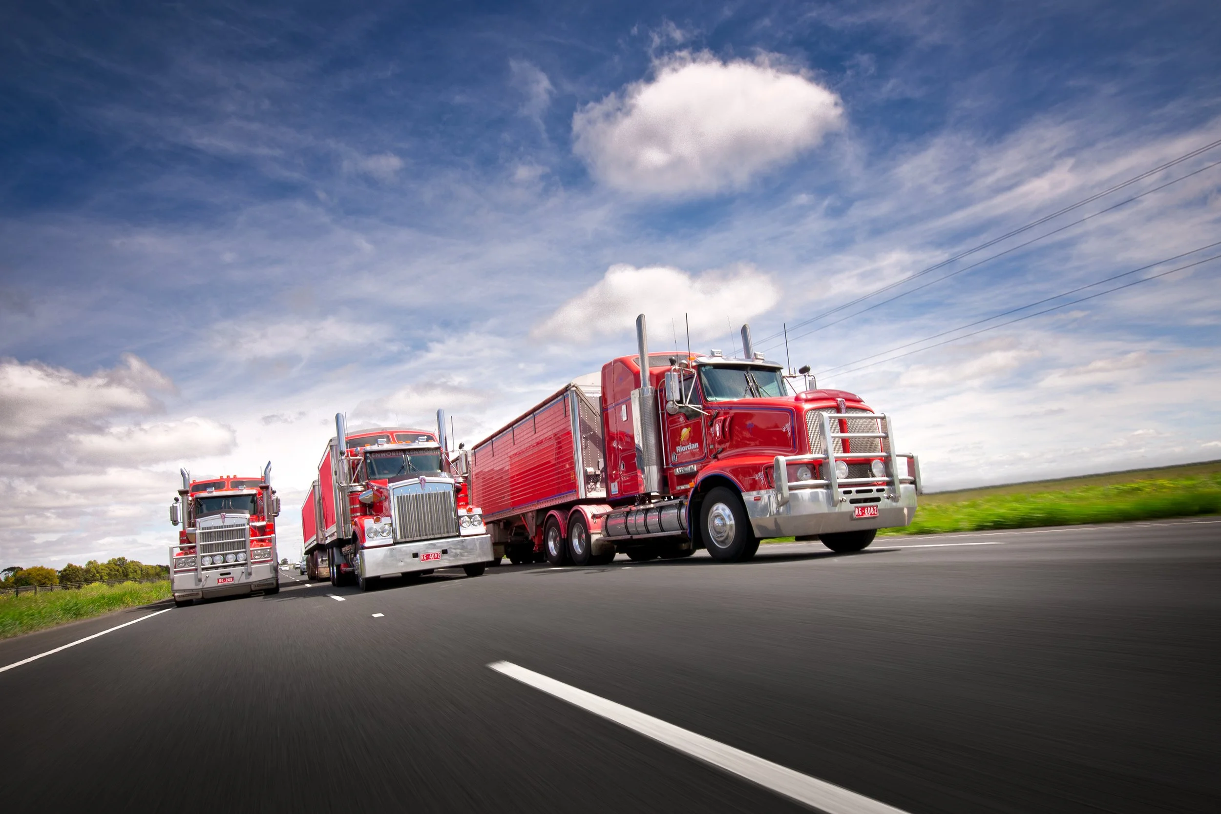 Three red fire trucks driving on a highway under a partly cloudy sky.