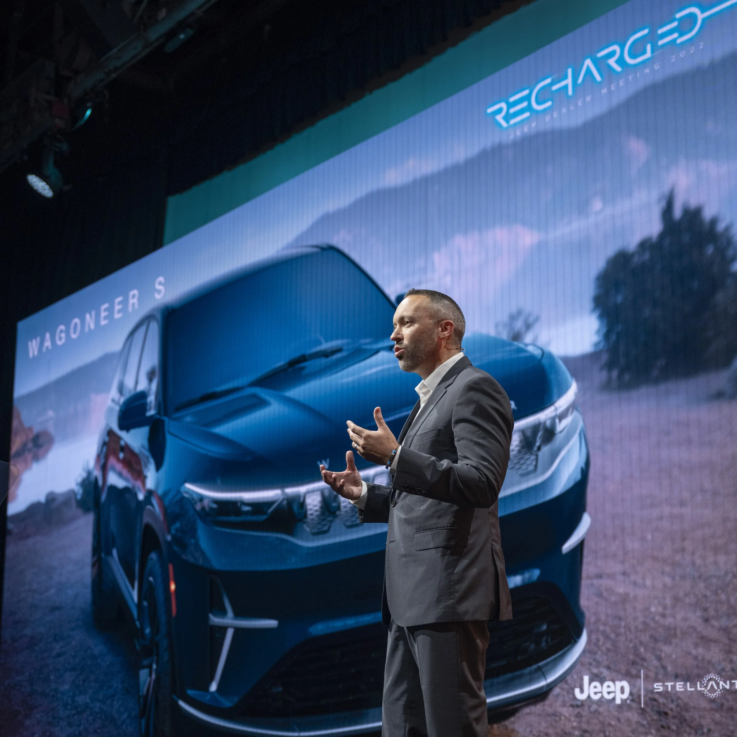 A man in a suit giving a presentation on stage with a large screen behind him displaying a vehicle and the words 'Wagoneer S' and 'Recharged.' The screen also shows a picture of a blue Jeep Wagoneer S SUV outdoors.