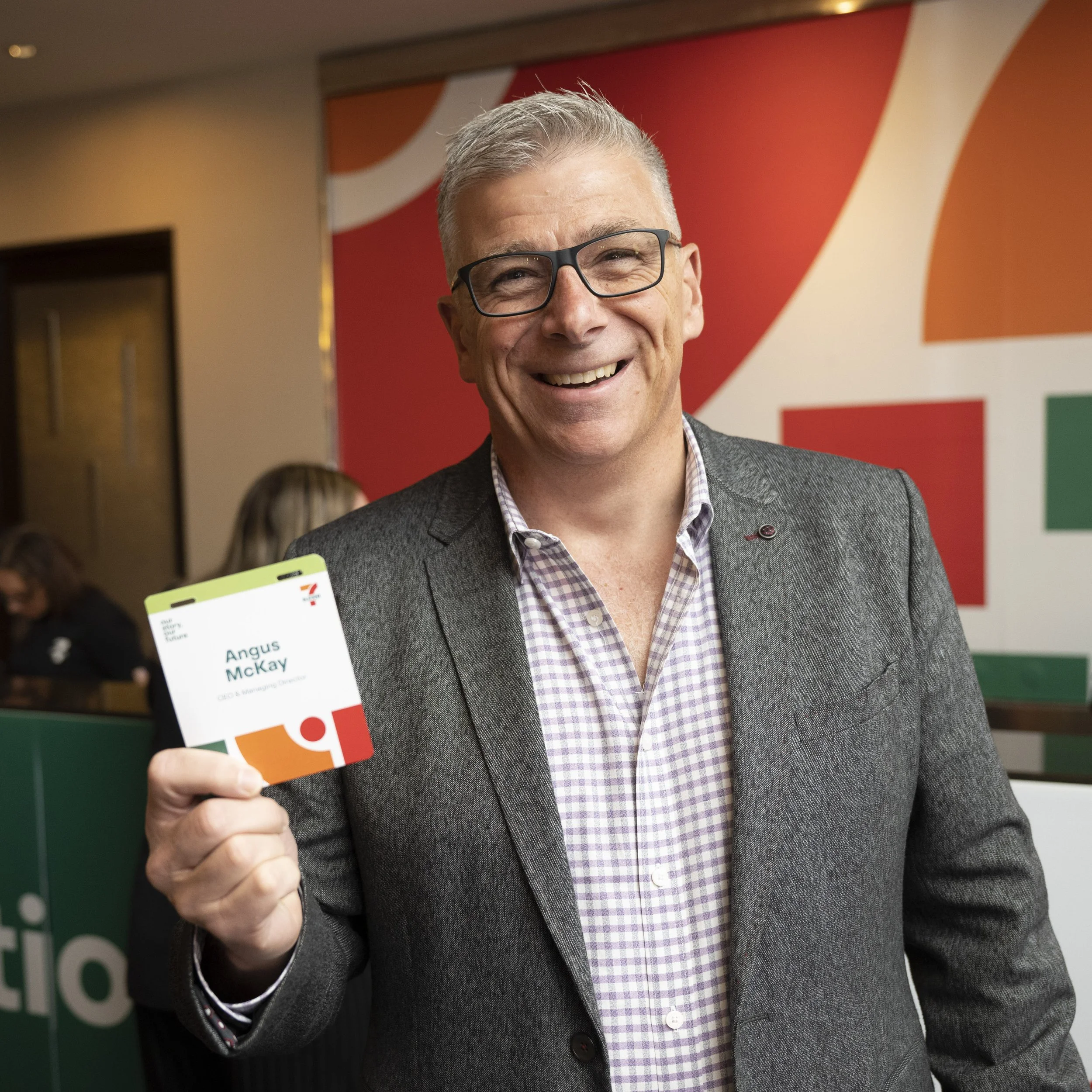 A smiling man wearing glasses and a gray blazer is holding a name badge that reads 'Angus McKay' at an event.