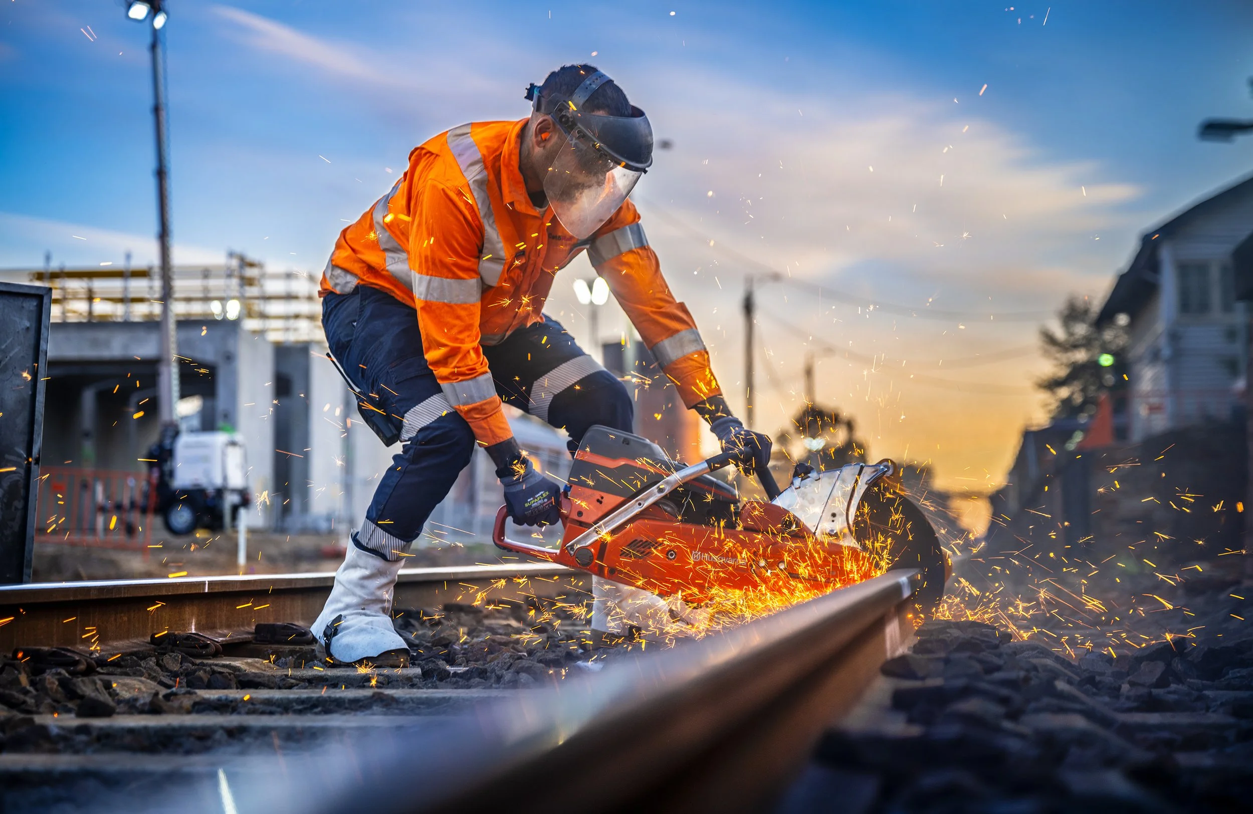 Worker cutting railroad track with a power saw, sparks flying, at sunset.