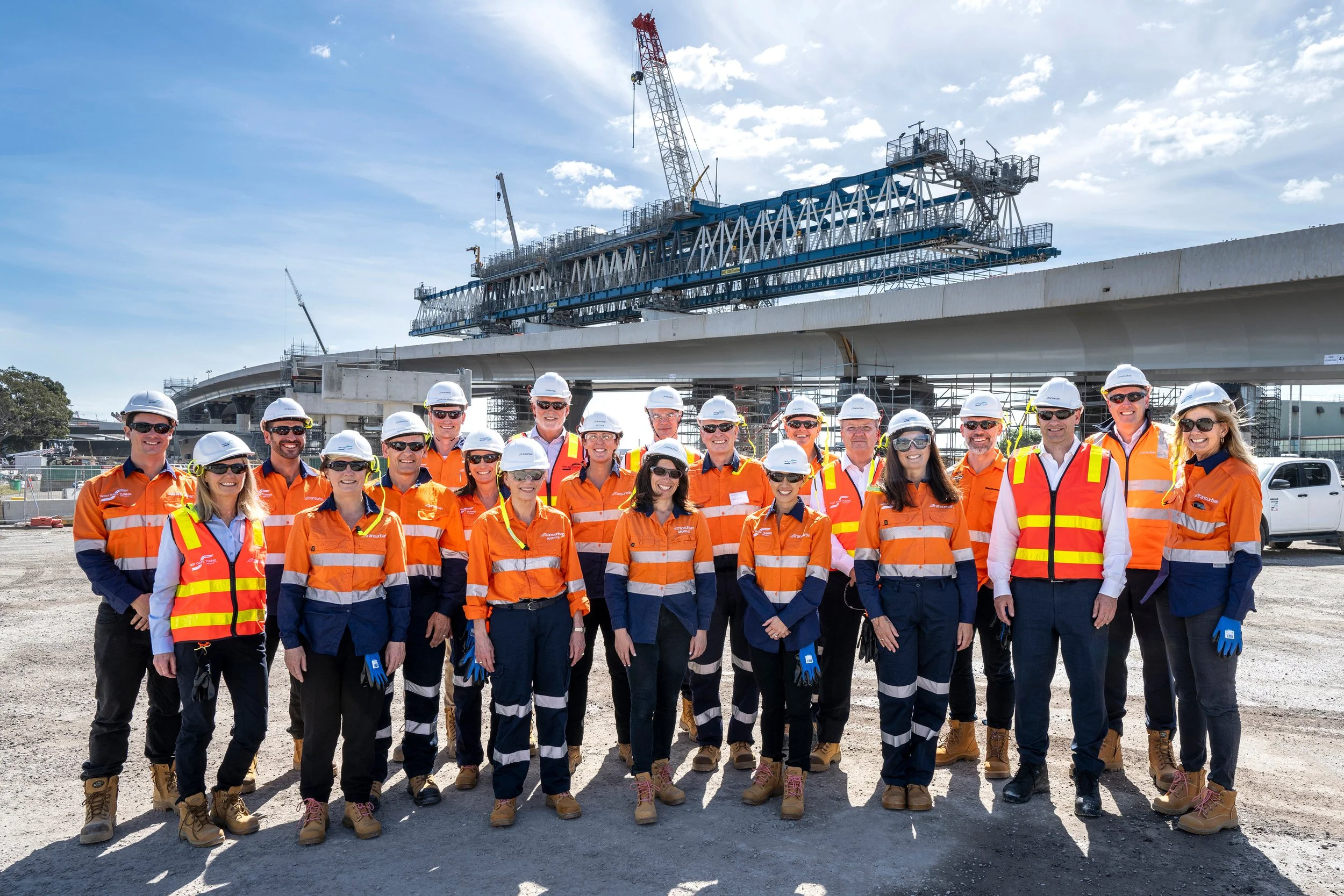 Group of construction workers in safety gear posing in front of a bridge construction site with cranes and scaffoldings, under a partly cloudy sky.