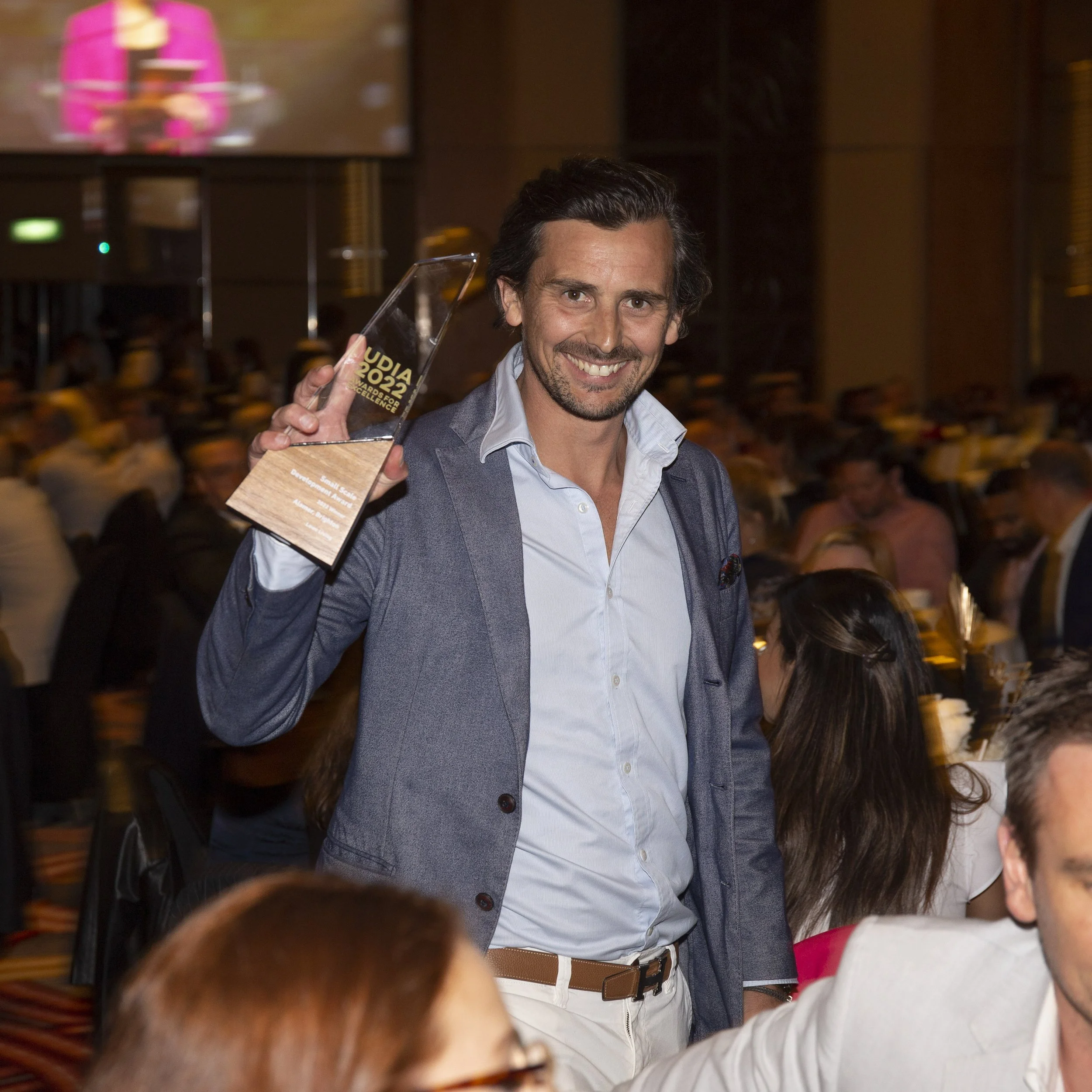 A man smiling and holding an award at a formal event, with many people seated around him in a banquet hall.