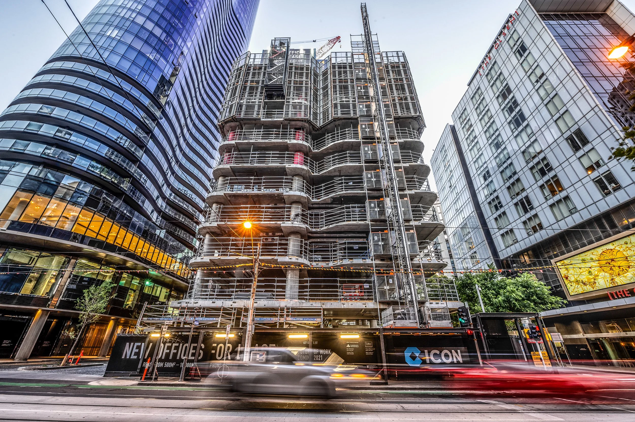 Under construction high-rise building on city street during twilight with other tall buildings surrounding it, moving vehicles in foreground and street lights