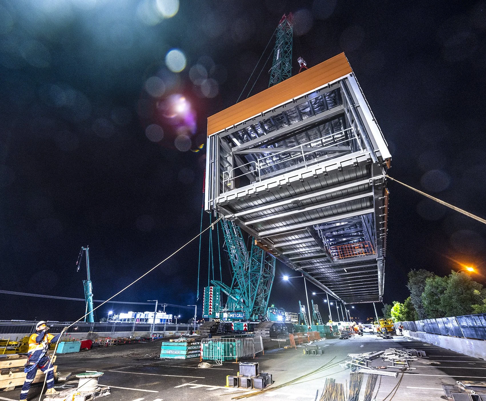 A construction worker wearing a safety vest and helmet stands on a construction site at night. Large construction crane lifts a massive container-shaped structure, illuminated under bright lights. The site is surrounded by fencing and equipment, with
