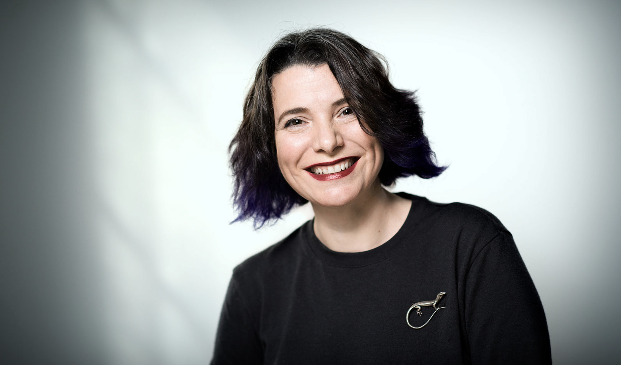 A smiling woman with shoulder-length dark hair with purple tips, wearing a black top and a silver lizard brooch, against a gray gradient background.