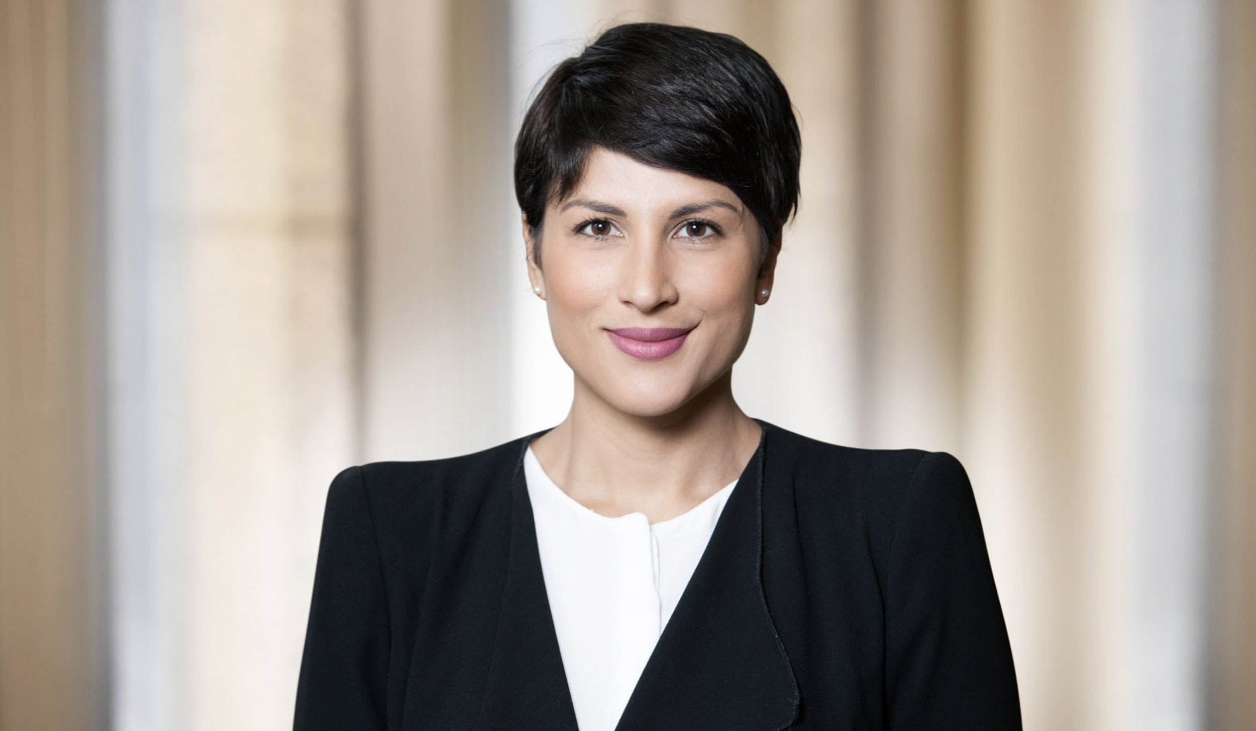A professional woman with short dark hair, wearing a black blazer and white top, smiling with a blurred background.