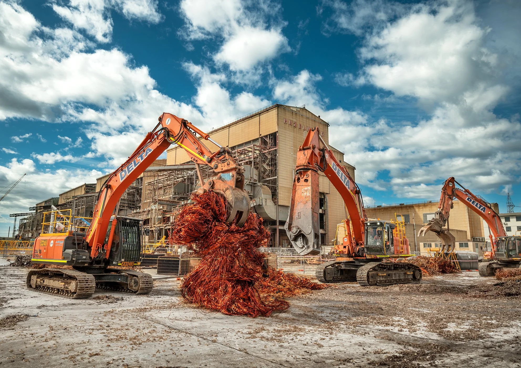 Construction site with three large excavators, one lifting tangled copper wire, in front of an industrial building under a partly cloudy sky.
