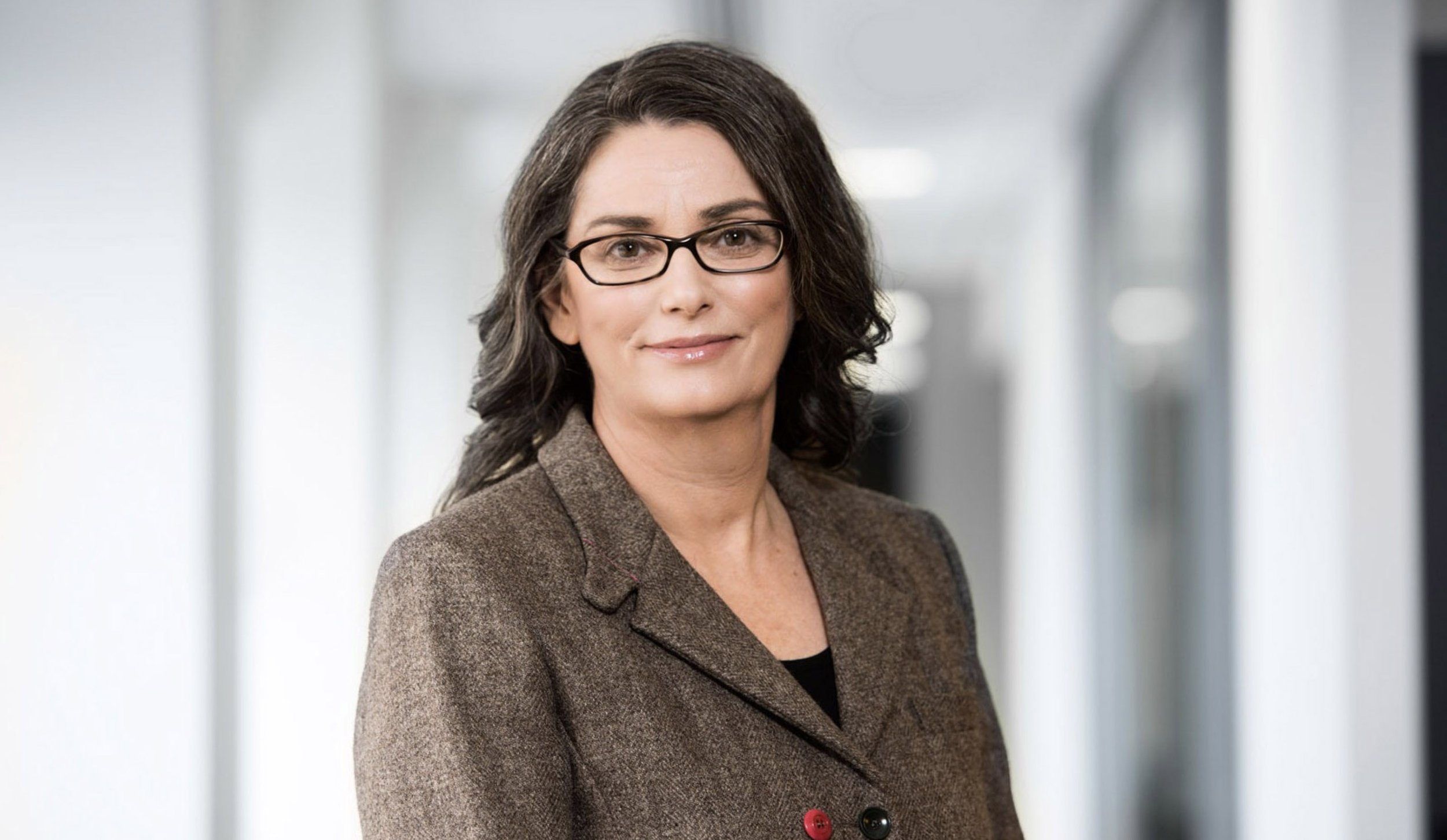 A professional woman with gray hair, glasses, and a brown blazer smiling in a corporate office corridor.