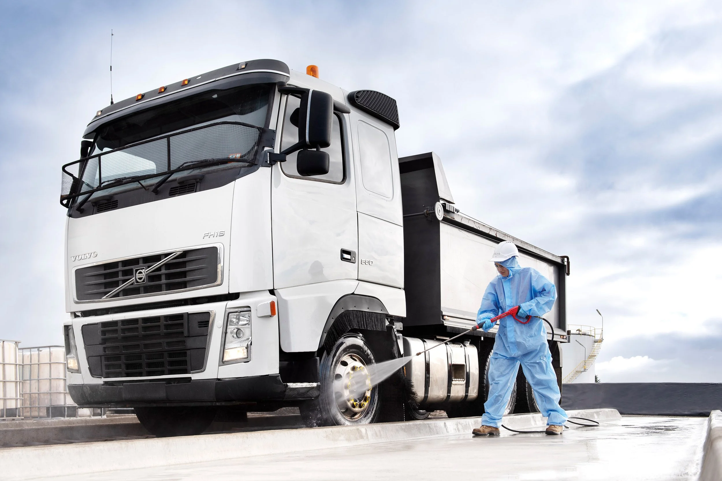 A worker in blue protective suit, mask, and gloves is pressure washing the tire of a large white truck on a rooftop or concrete surface.