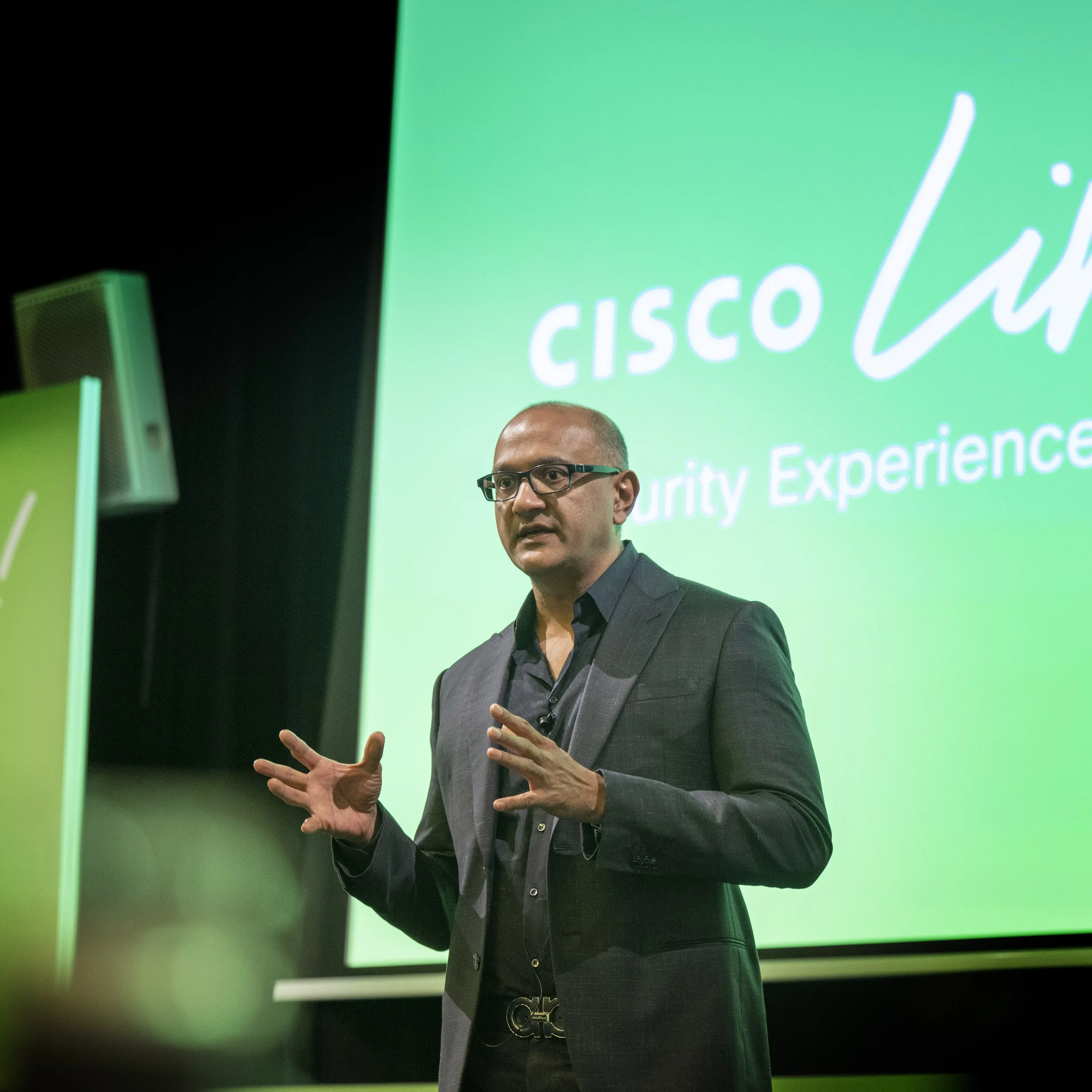 A man in a dark suit and glasses speaking at a Cisco security conference, with a large green Cisco Security logo projected behind him.