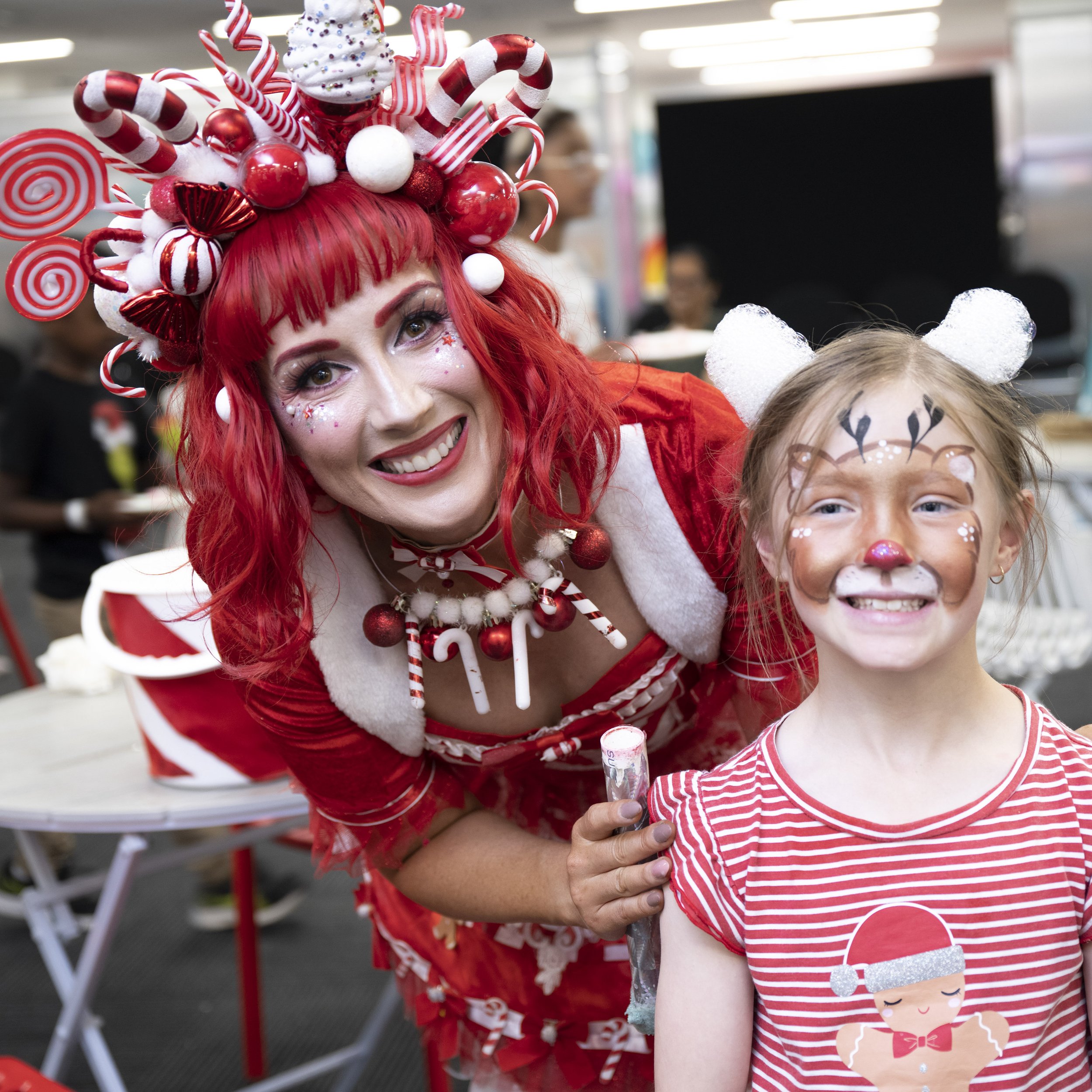 A woman dressed in a Christmas-themed costume with red hair and a festive headpiece stands next to a young girl with face paint resembling a reindeer, both smiling at the camera.