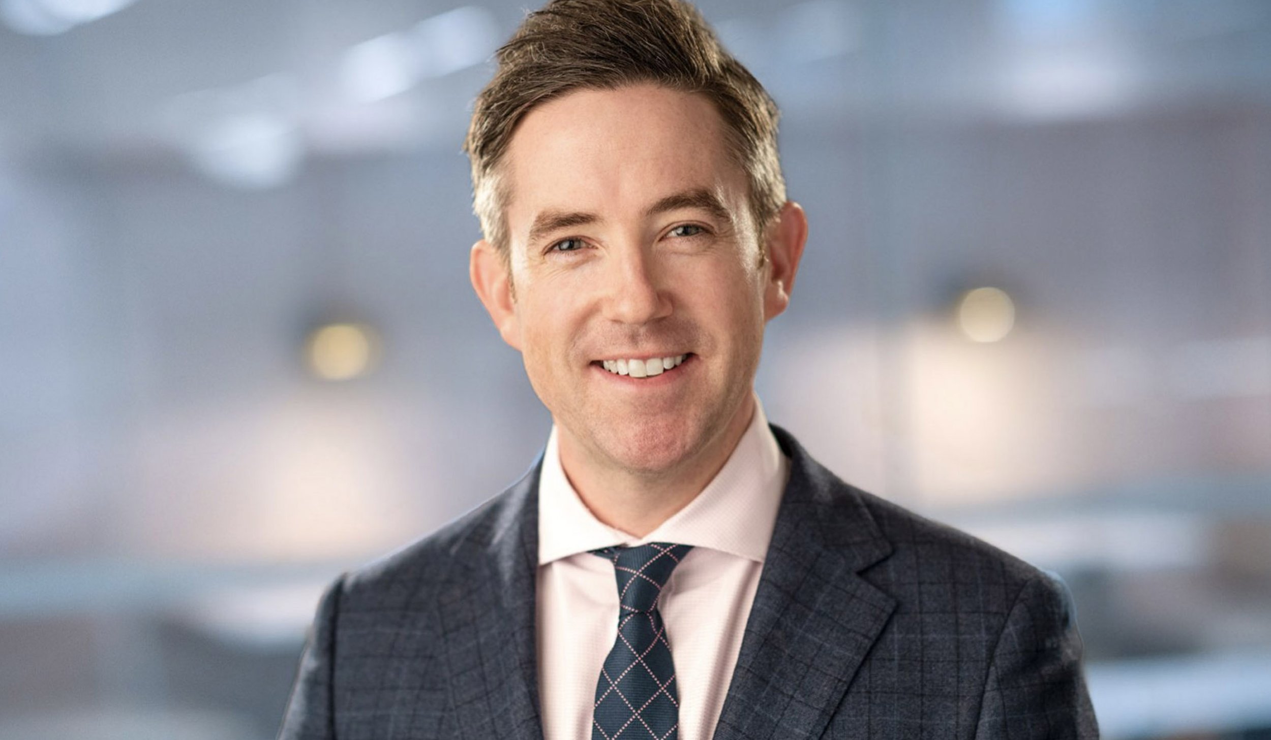 A smiling man in a suit and tie standing in an indoor setting with blurred background.