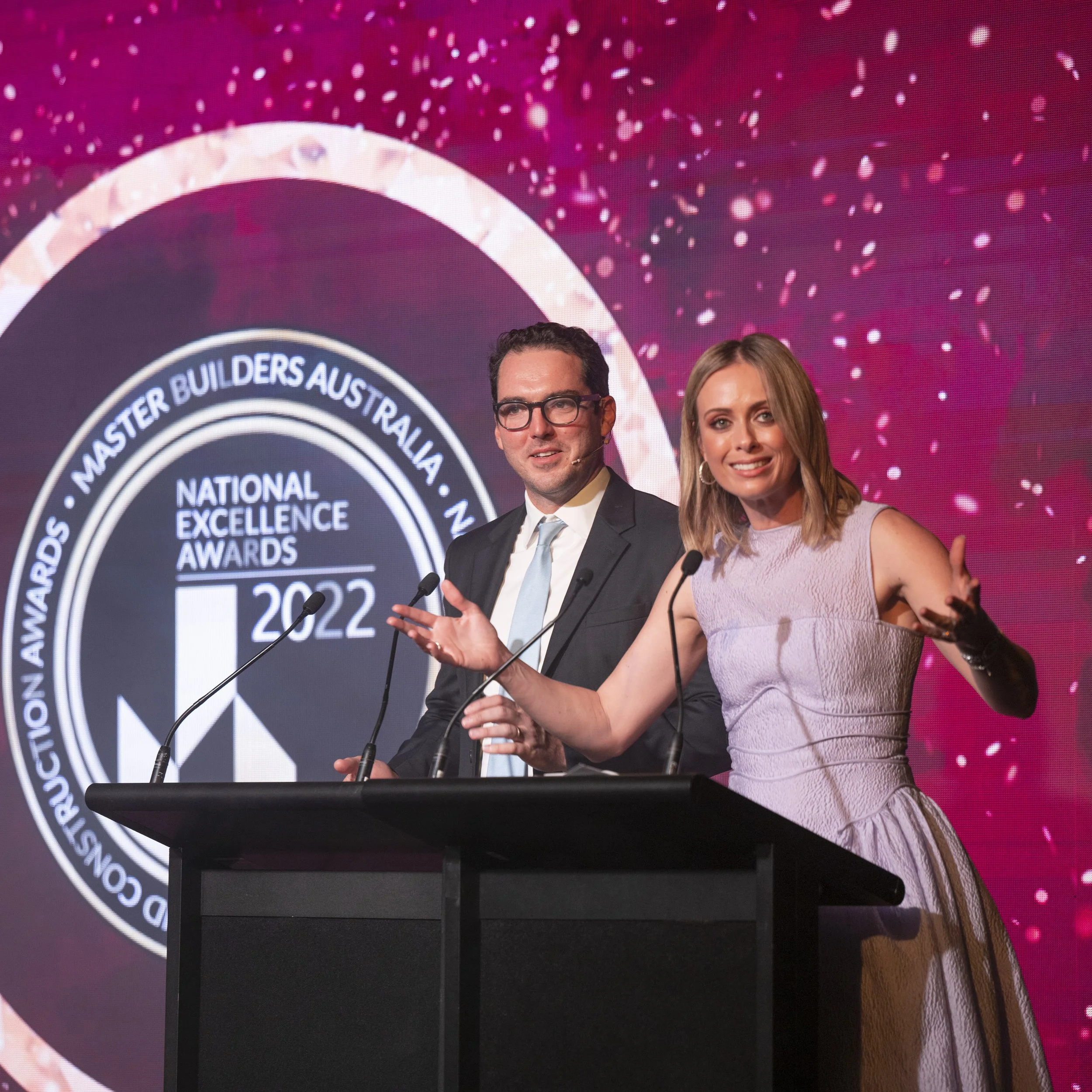 Two people, a man in a dark suit and glasses and a woman in a sleeveless light purple dress, speaking at a podium during the 2022 National Excellence Awards, with a large pink and black logo behind them.
