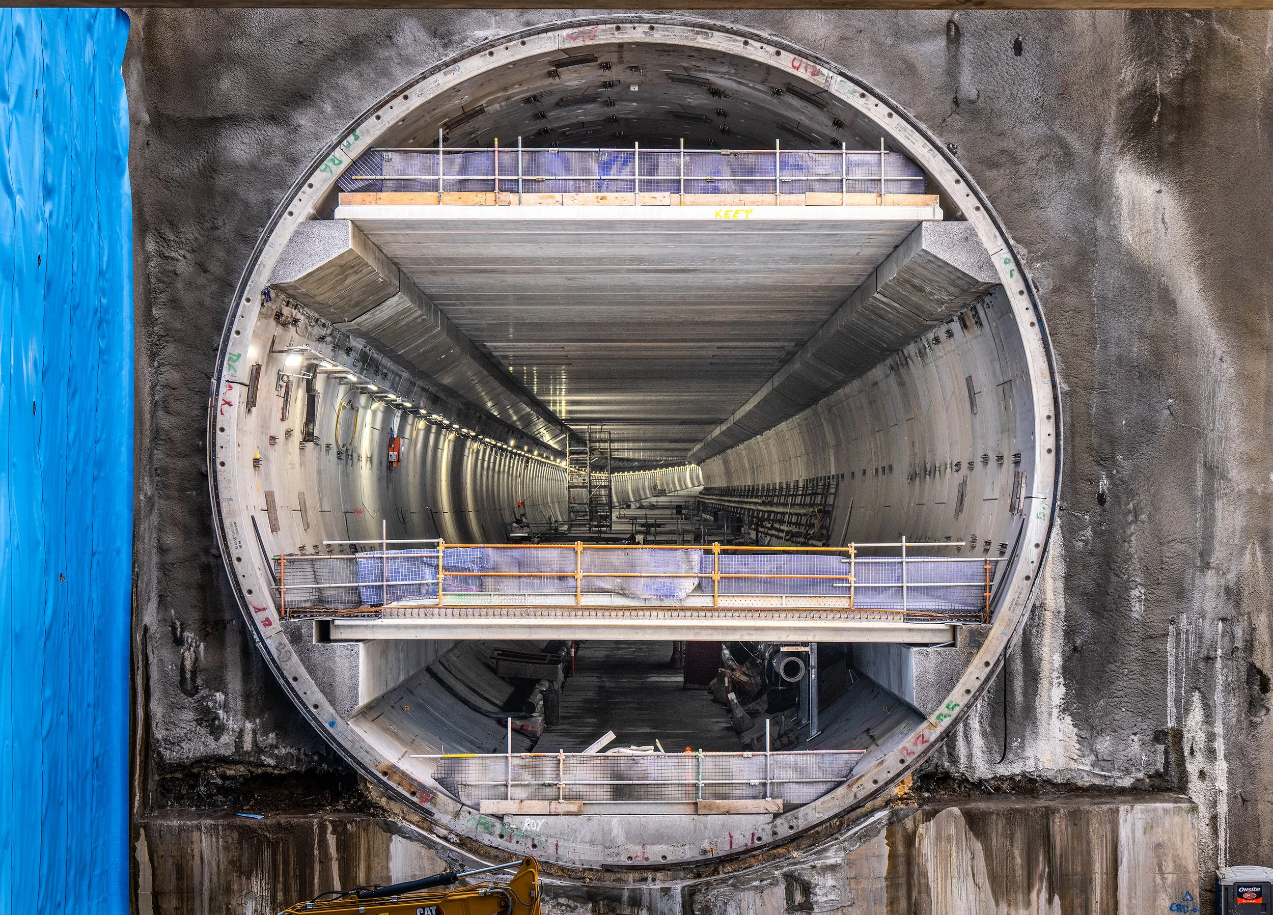 Construction scene inside a large tunnel under construction with scaffolding and lighting, viewed through an excavated circular wall.