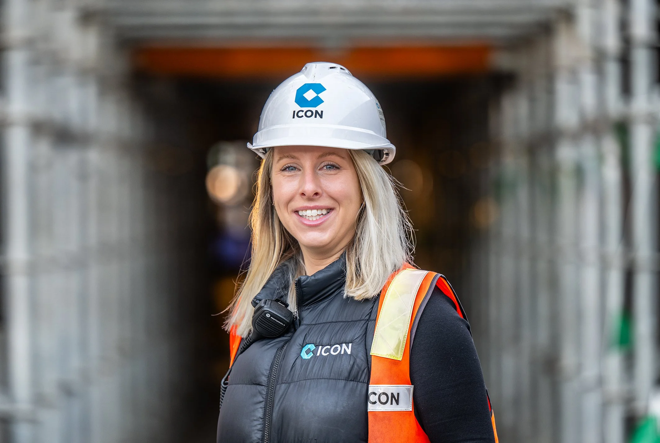 A smiling woman wearing a white hard hat with an ICON logo, a black vest with the ICON logo, and an orange safety vest, standing in a construction area.