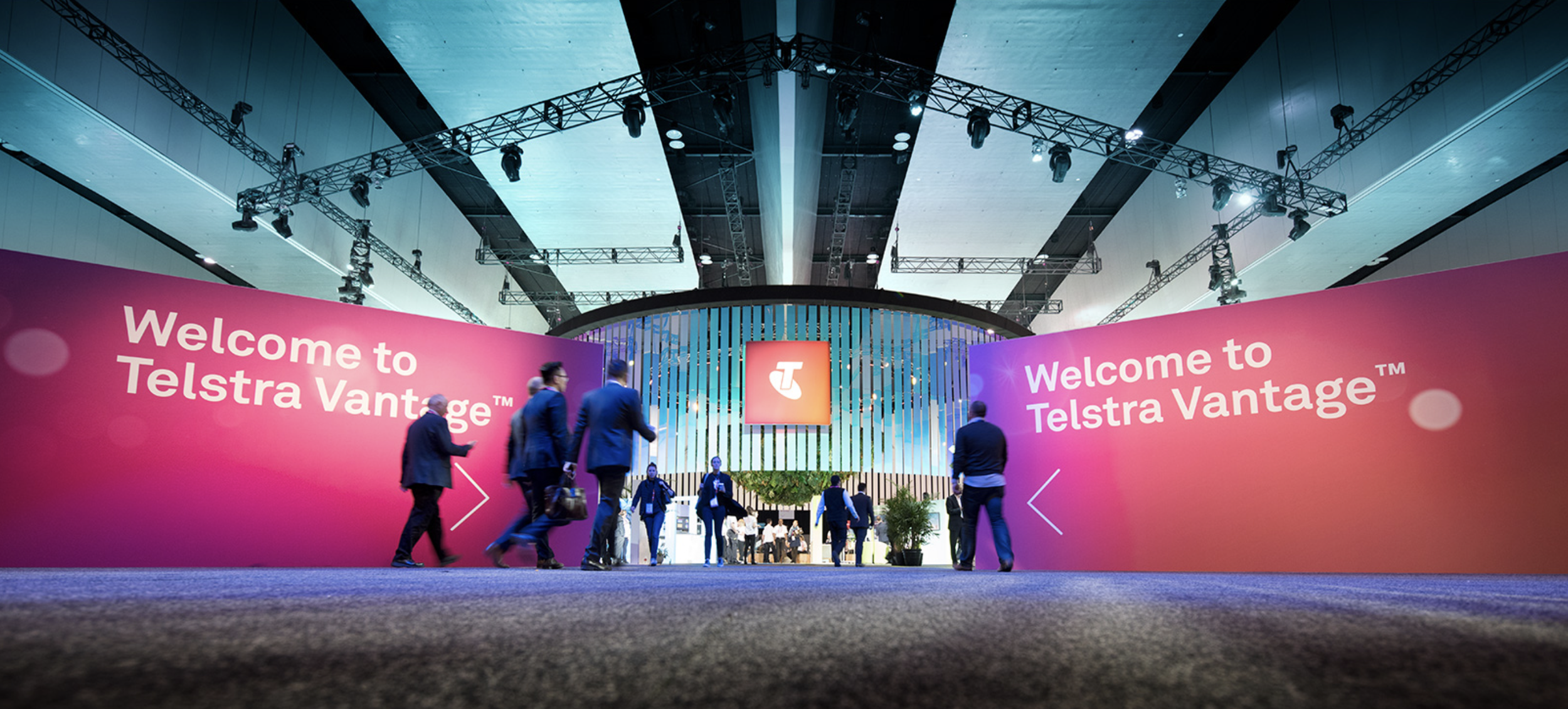 Indoor conference or trade show with a large pink and purple welcoming banner reading 'Welcome to Telstra Vantage.' People in business attire walking and conversing in front of booths and displays, with an overhead lighting rig and a central digital 