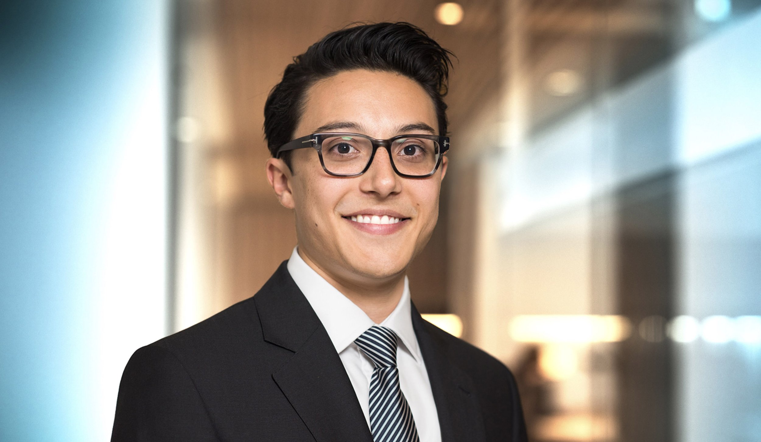 Headshot of a young man with dark hair, glasses, wearing a black suit, white shirt, and striped tie, smiling in a modern office environment.