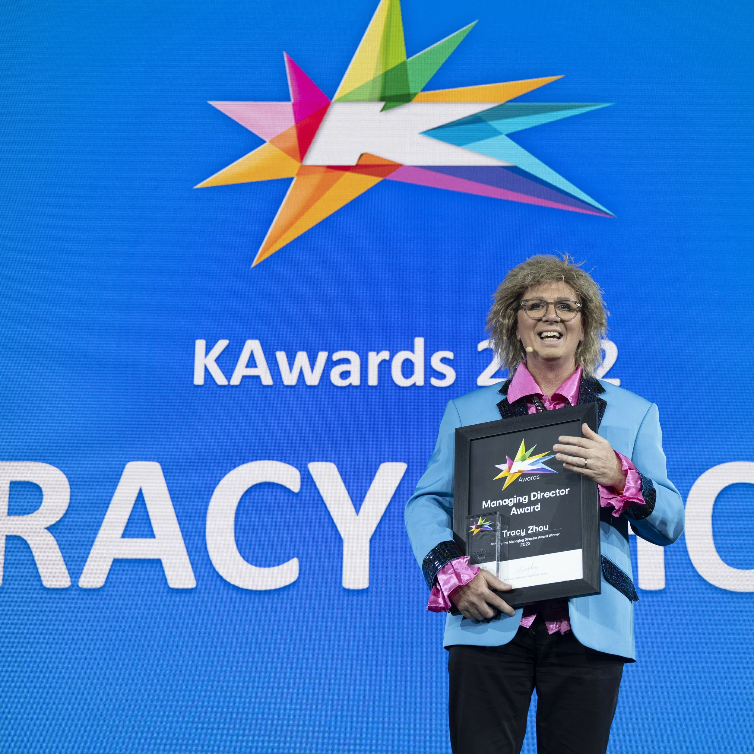 Person wearing colorful blazer holding a framed award on stage at a conference with a large screen displaying a star logo and text in the background.