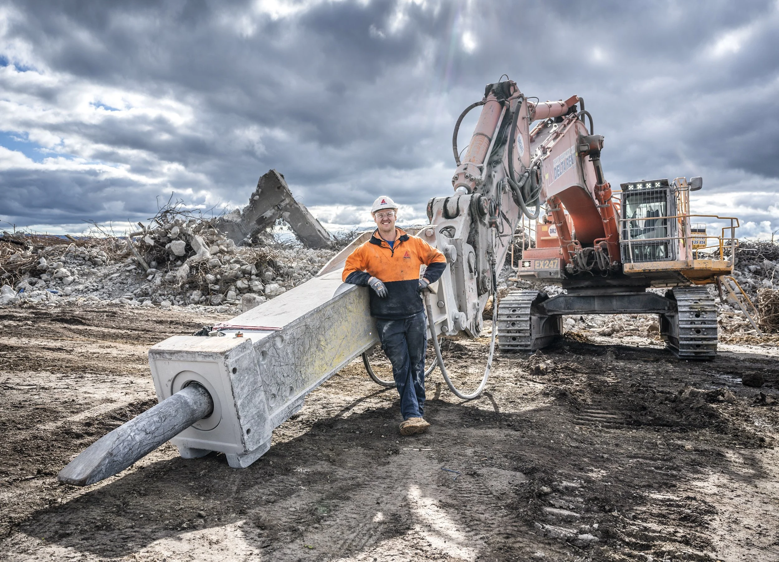 A construction worker in an orange and black jacket stands next to large machinery on a dirt site with debris and rubble, under a cloudy sky.