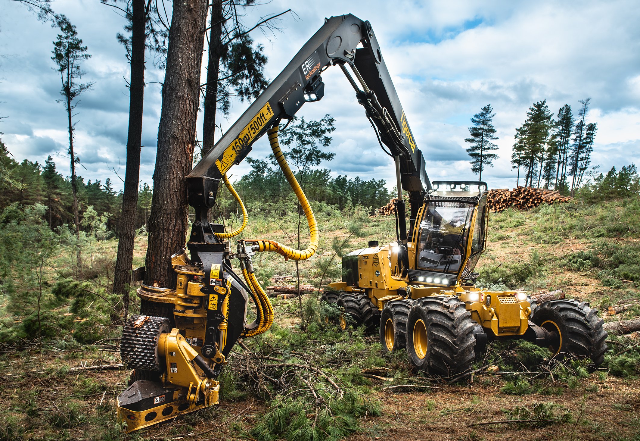 A yellow forestry mulcher with a long arm attachment is working in a forest, clearing small trees and brush. The machine is on rugged terrain with tall trees and a cloudy sky in the background.