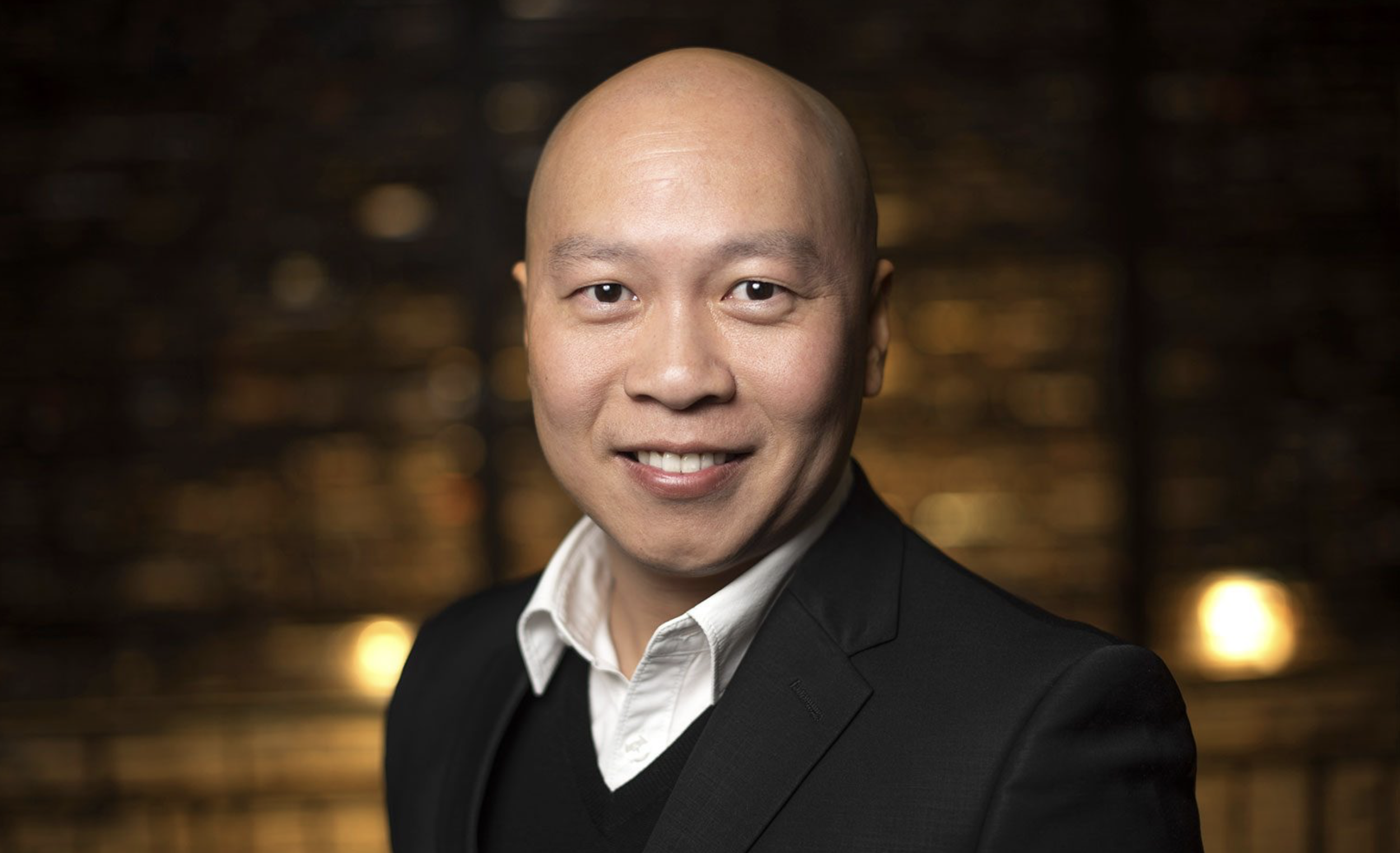 A smiling Asian man with a shaved head in business attire, against a dark, blurred background with warm lighting.