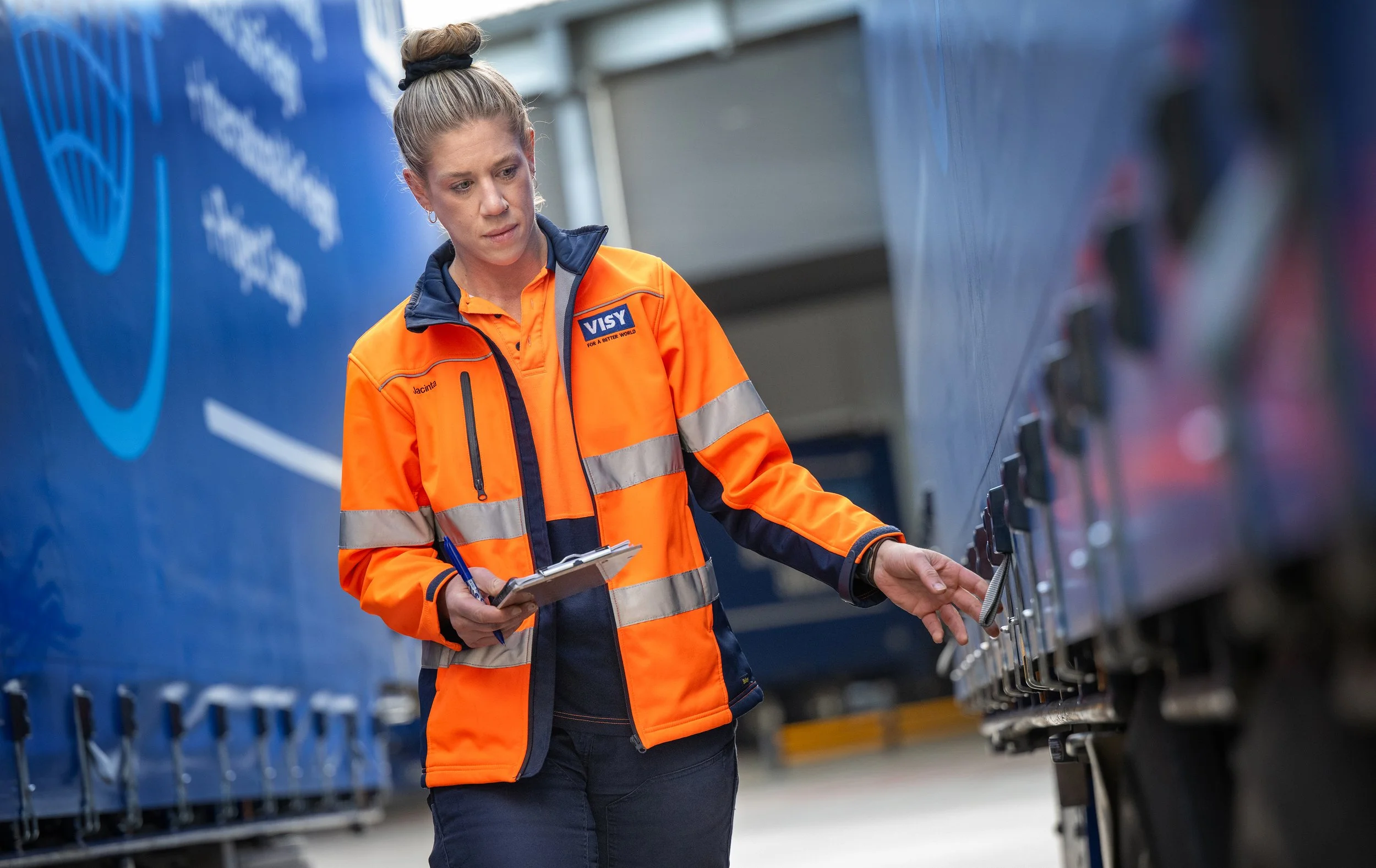 A woman in an orange reflective safety jacket with a clipboard inspecting a truck's security latch inside a warehouse or distribution center.