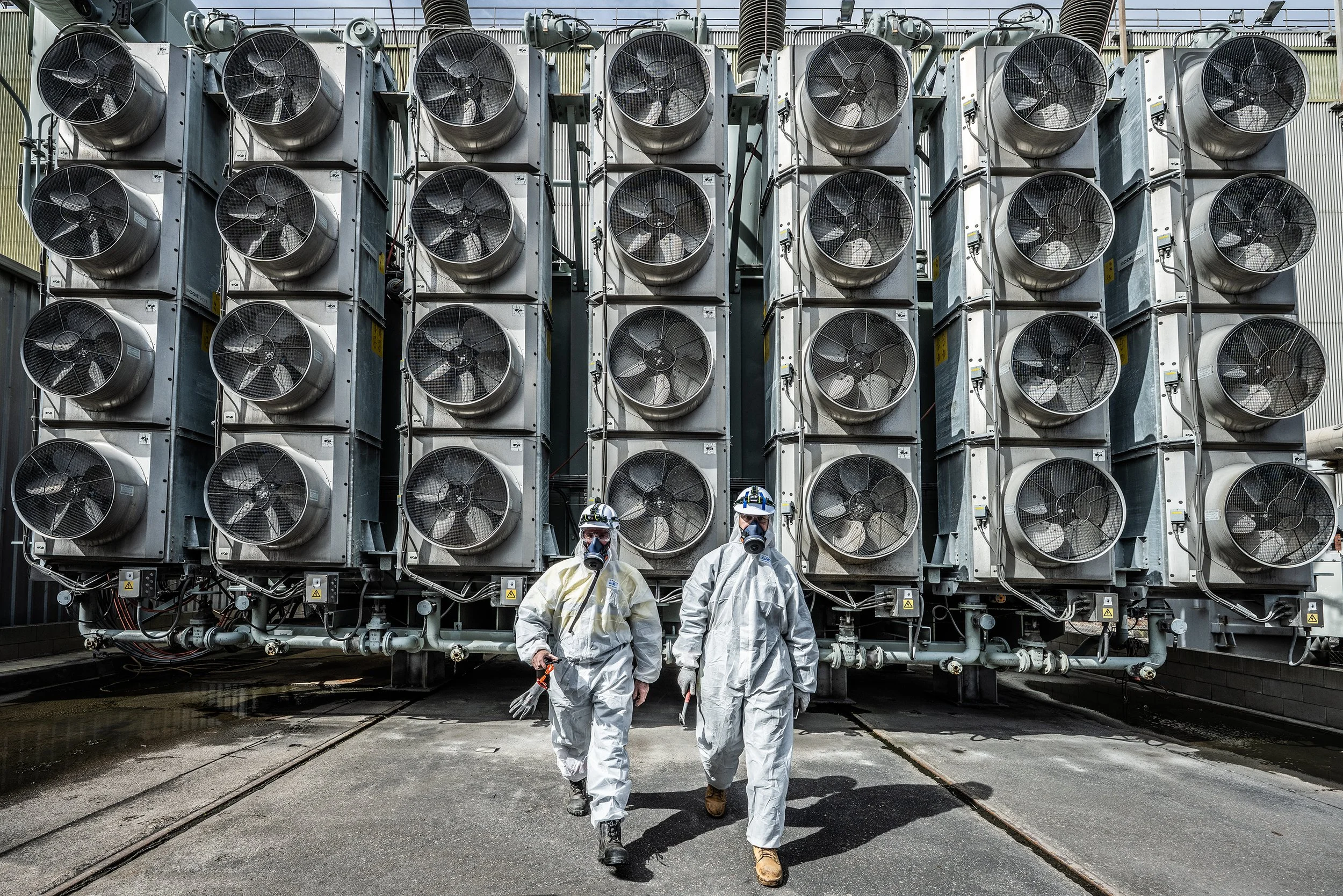 Two workers in protective suits and masks walking in front of a large industrial cooling system or heat exchanger with multiple fans.
