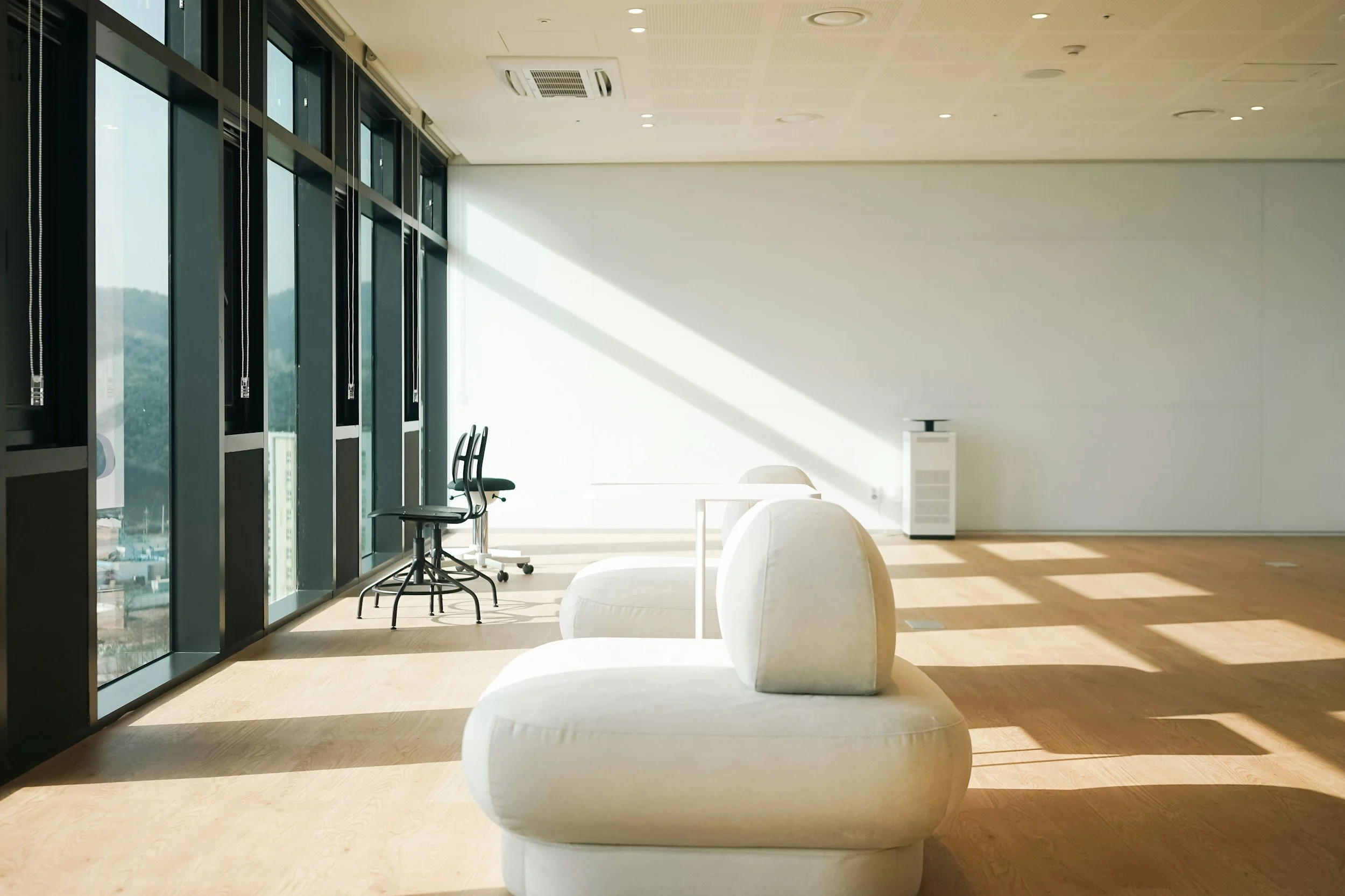 Modern office lounge area with white curved seating, large glass windows letting in sunlight, and a few chairs near the windows. The floor is wooden, and the ceiling has built-in lighting and an air conditioning vent.