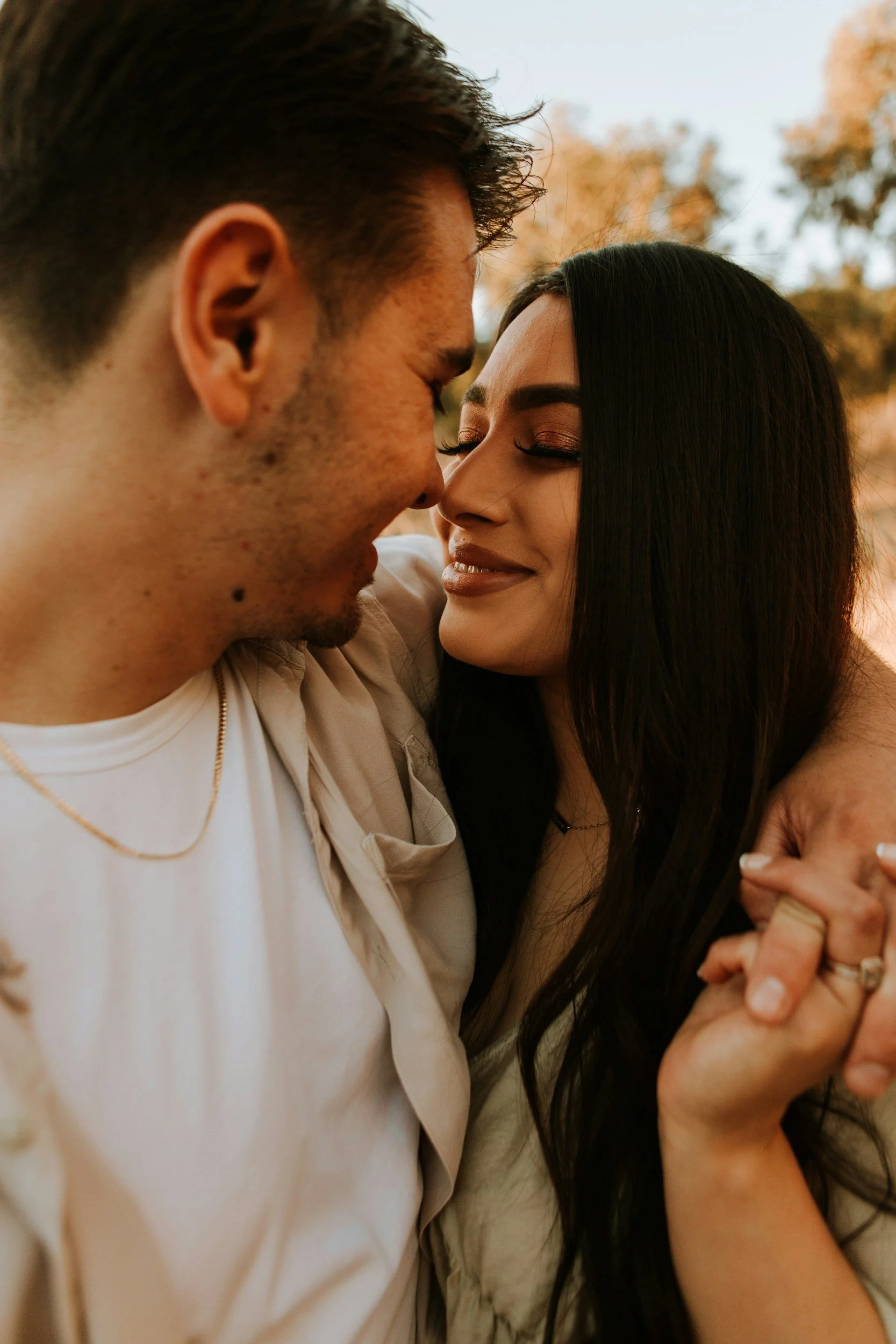 A man and a woman are close, smiling, with foreheads and noses touching, outdoors during sunset, with trees in the background.