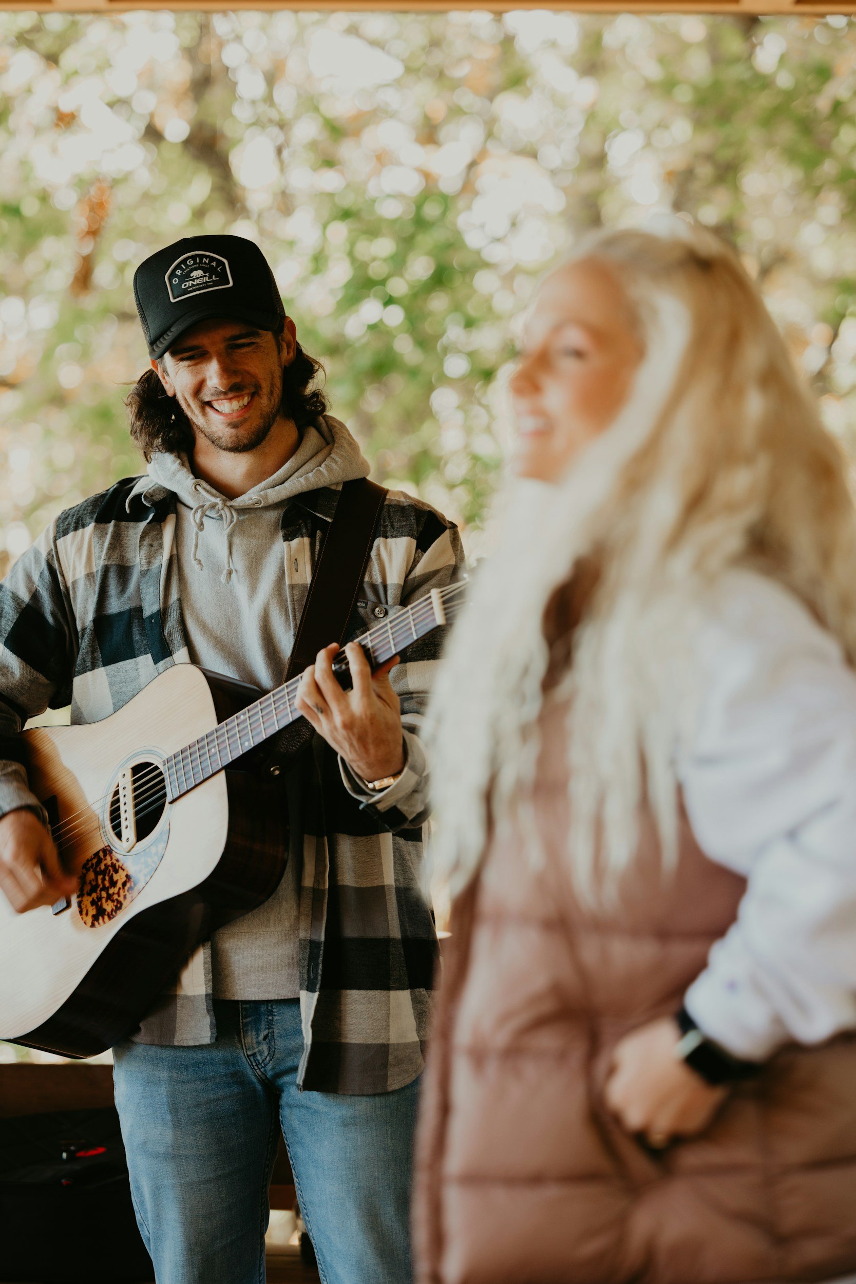 A man with long dark hair wearing a black cap, hoodie, and plaid shirt playing an acoustic guitar, smiling, standing next to a woman with long blonde hair in a cream-colored sweater, sitting at a table, laughing, with blurred trees in the background.