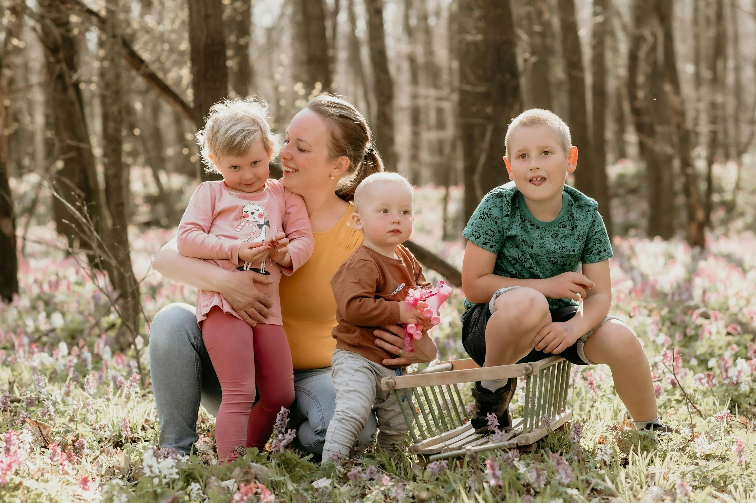 A woman and four children sitting outdoors in a natural setting with blooming flowers and trees, some sitting on a small wooden bench and one on her lap, enjoying a sunny day.