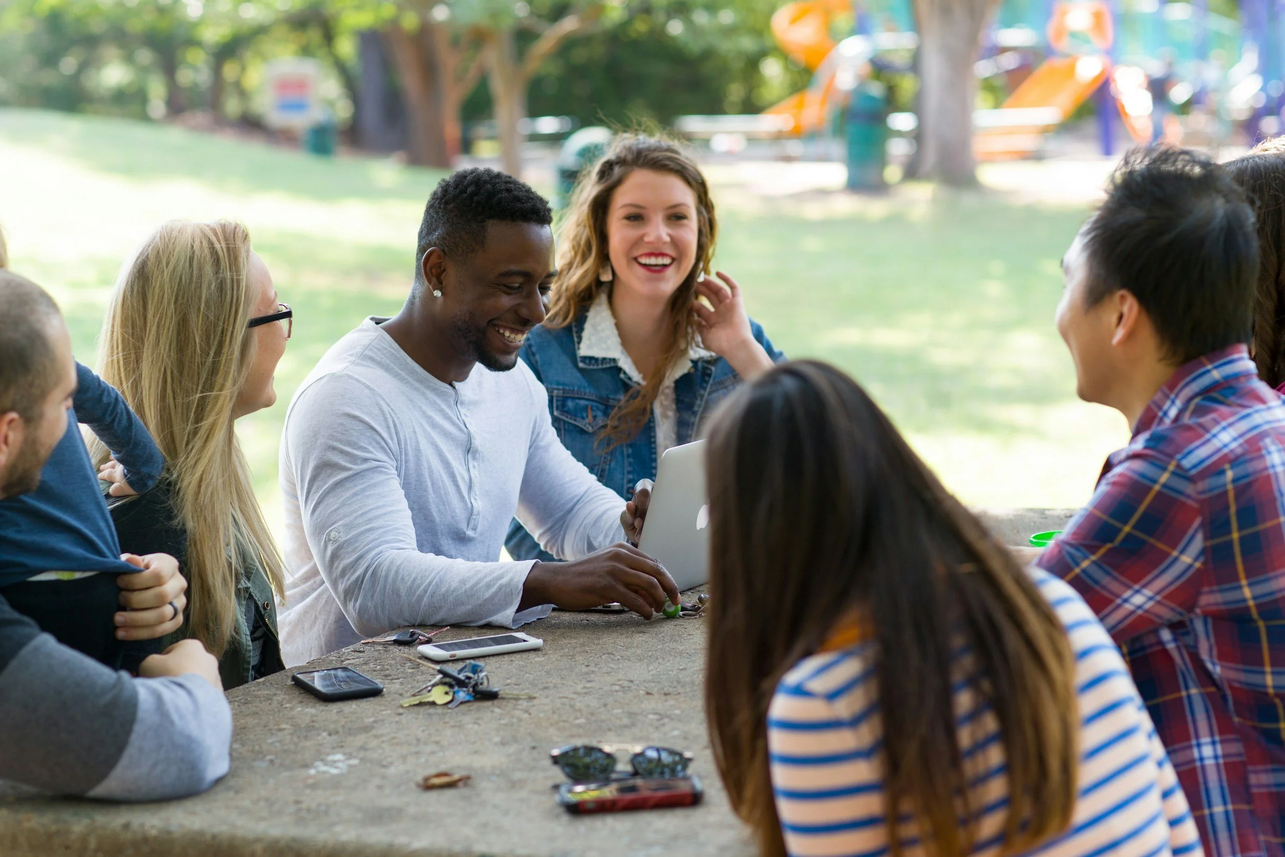 A group of people sitting outdoors at a park table, smiling and engaging with each other, with trees and playground in the background.