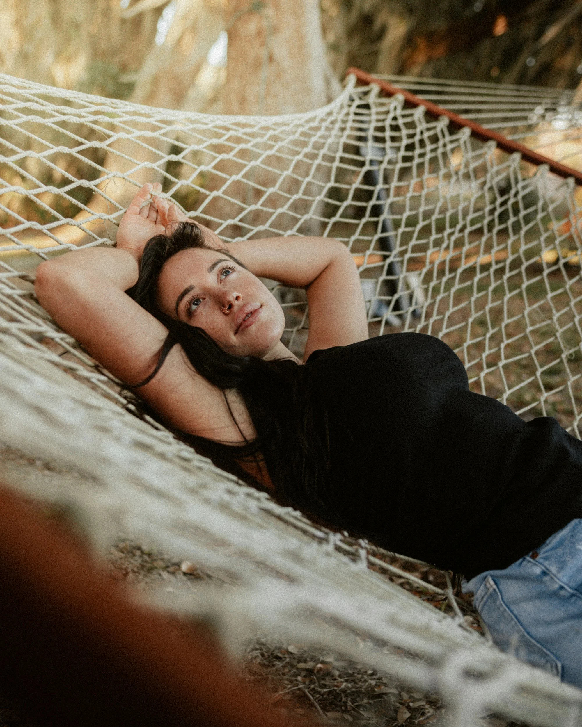 A woman lying in a hammock outdoors, looking upward and resting her head on her arm, with trees and leaves in the background.