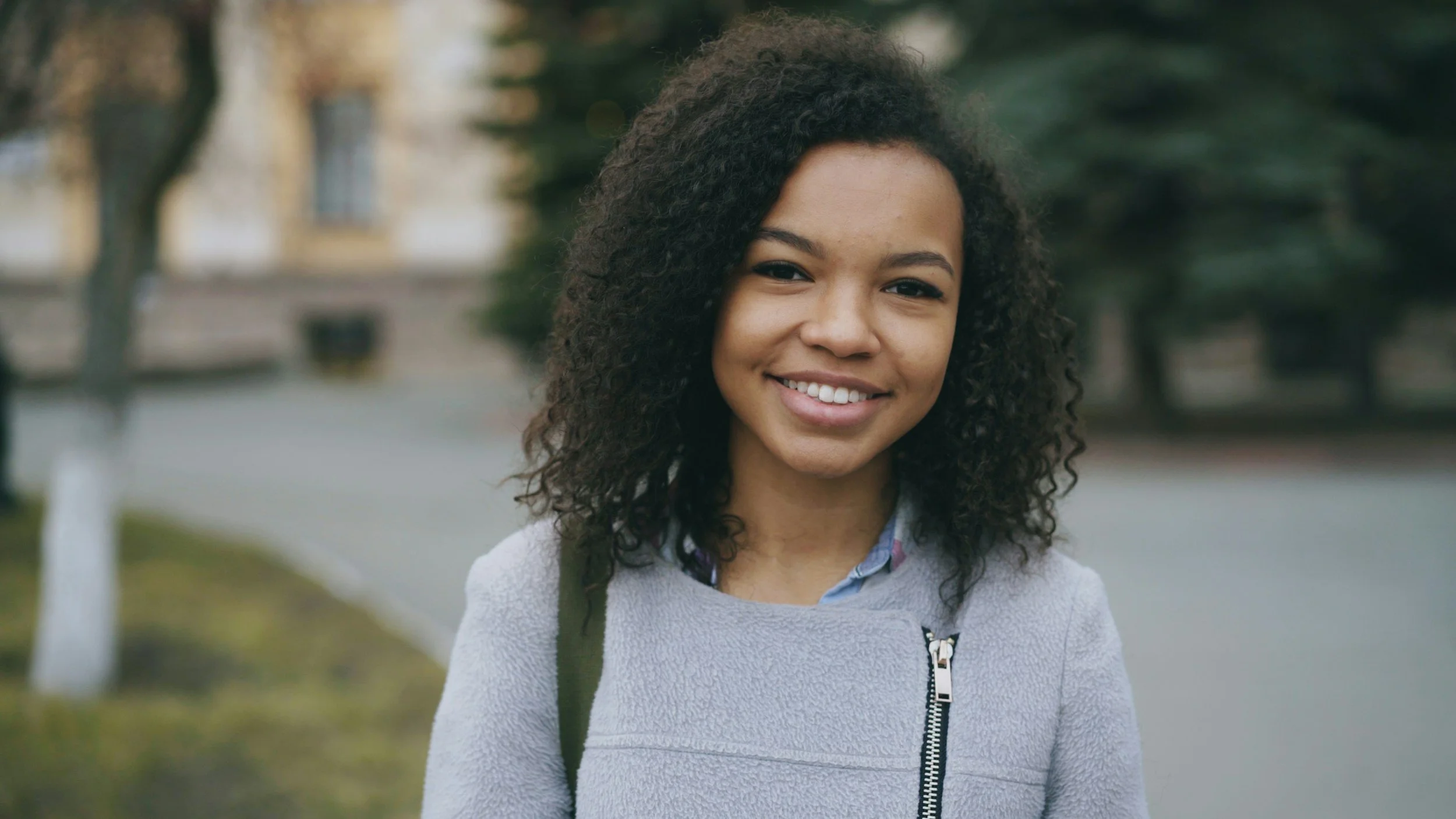 A young woman with curly hair smiling outdoors, wearing a light gray sweater and a floral shirt, with trees and a park path in the background.