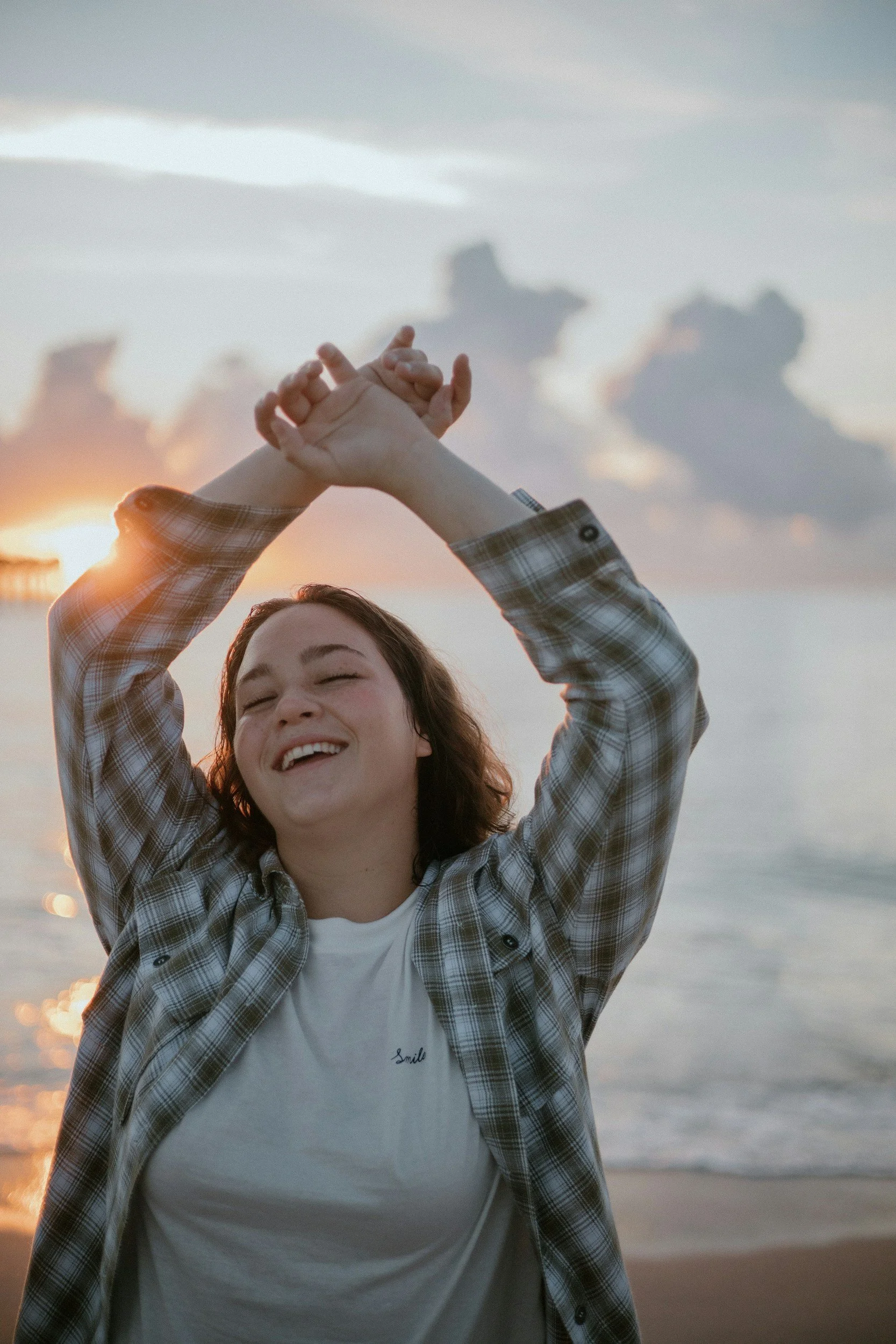 A smiling young woman with eyes closed, wearing a plaid shirt over a white t-shirt with the word 'Smile' embroidered, standing on a beach during sunset, with her arms raised and hands clasped above her head.